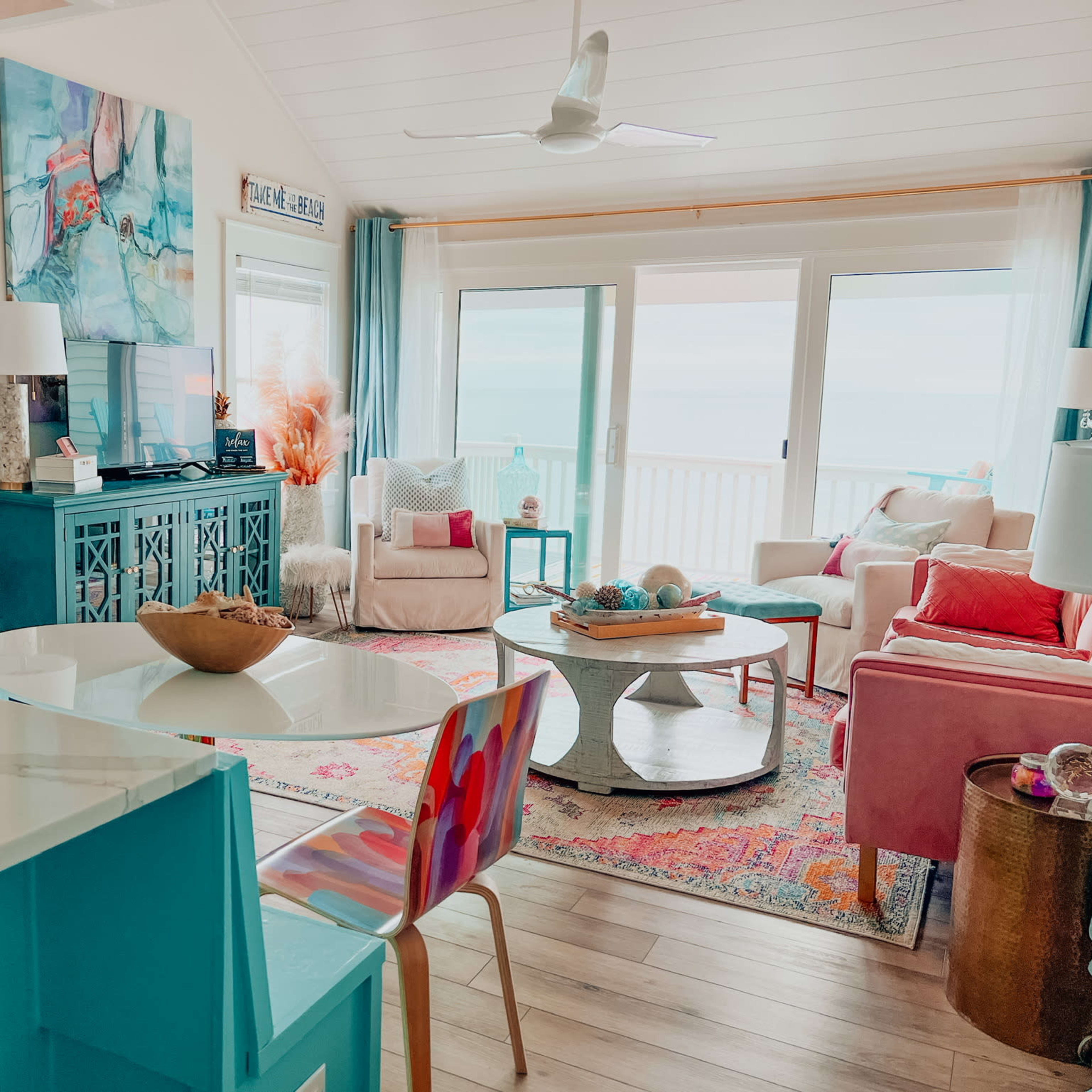 A bright living area with a turquoise cabinet, pink chairs, and a large window overlooking a beach view.