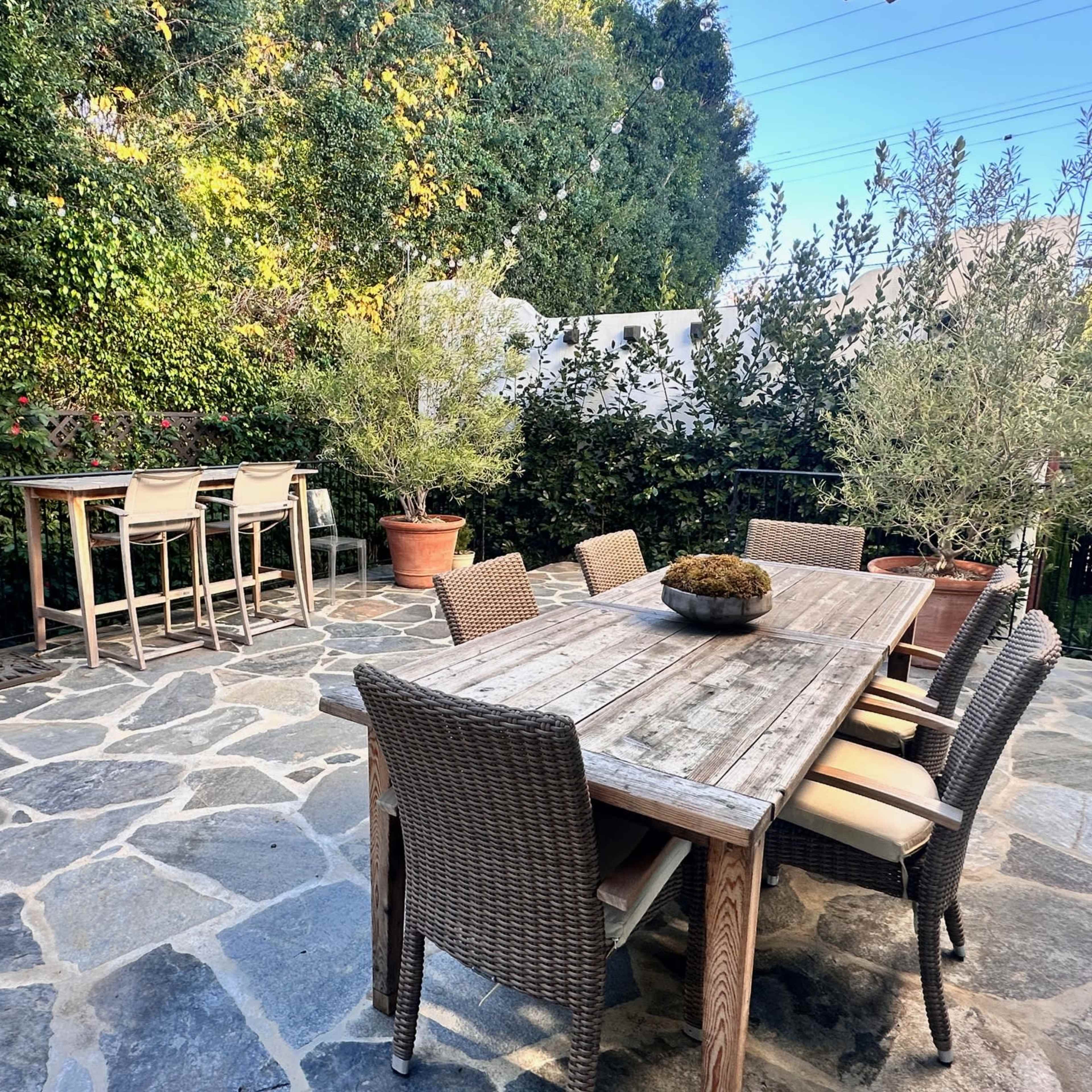 A patio features a large wooden table surrounded by wicker chairs, with a bar-height table and chairs set against a backdrop of greenery.