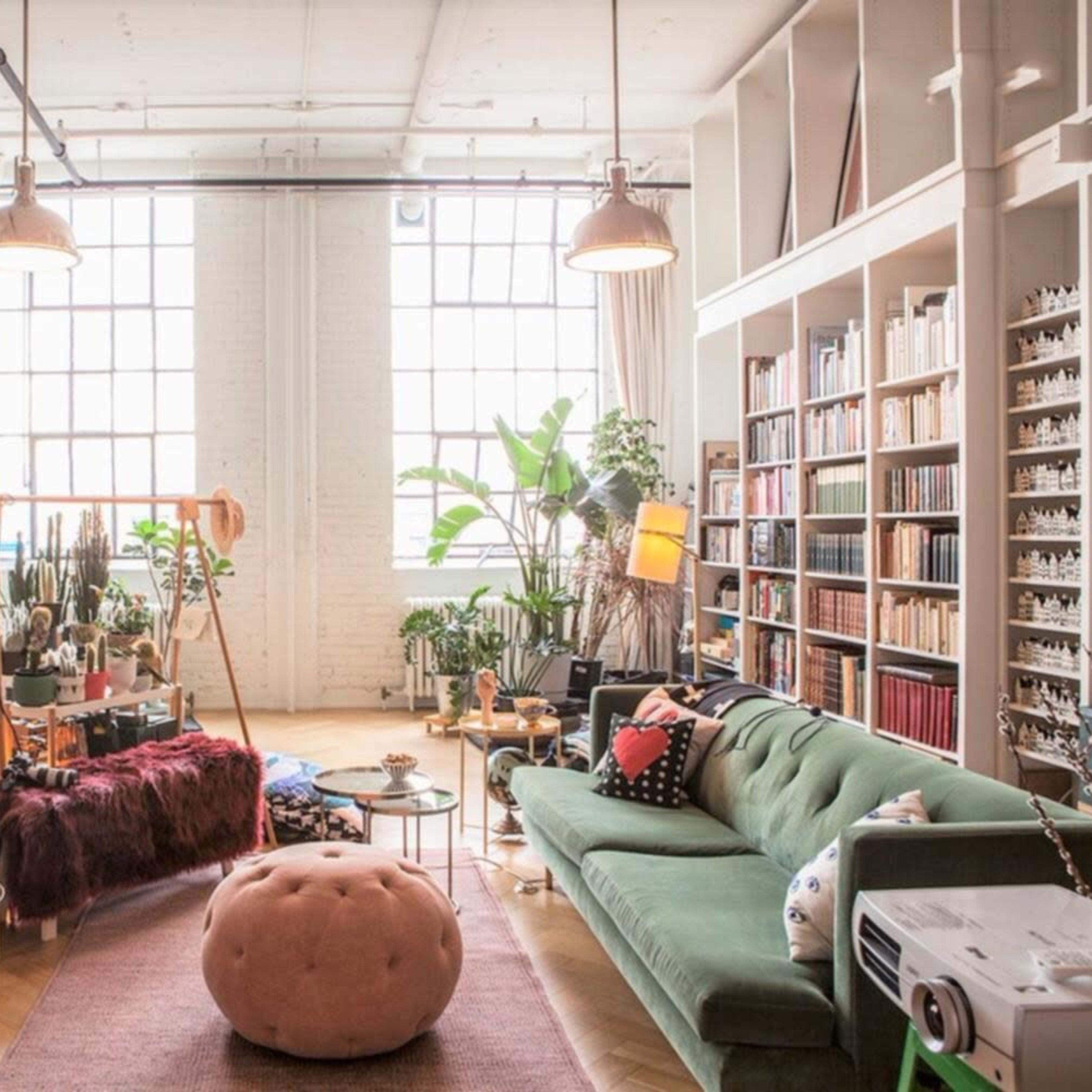 A spacious living room features large windows, bookshelves filled with colorful books, a green sofa, and various plants.