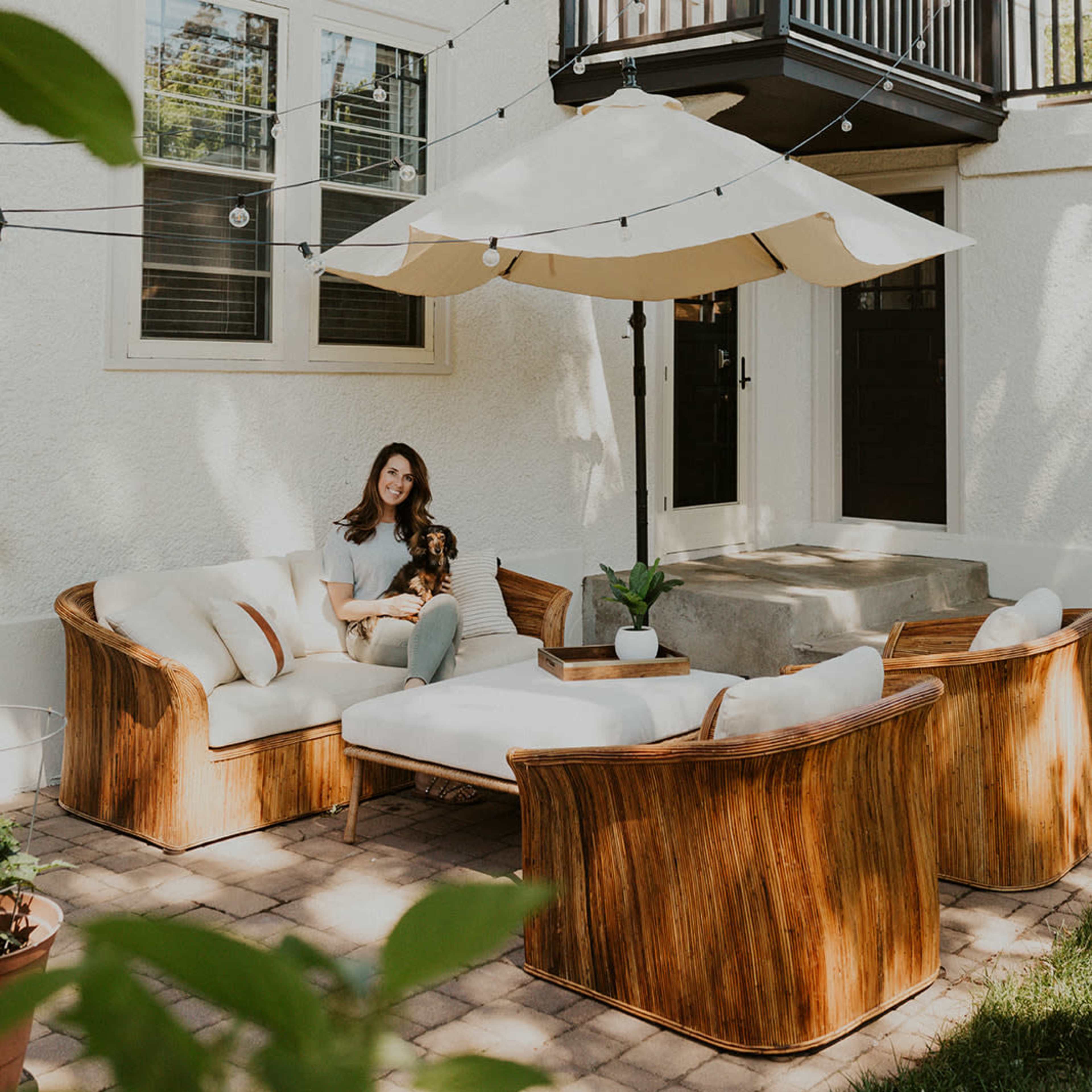 A woman sits on a wicker couch in a backyard area with a large umbrella and string lights above.