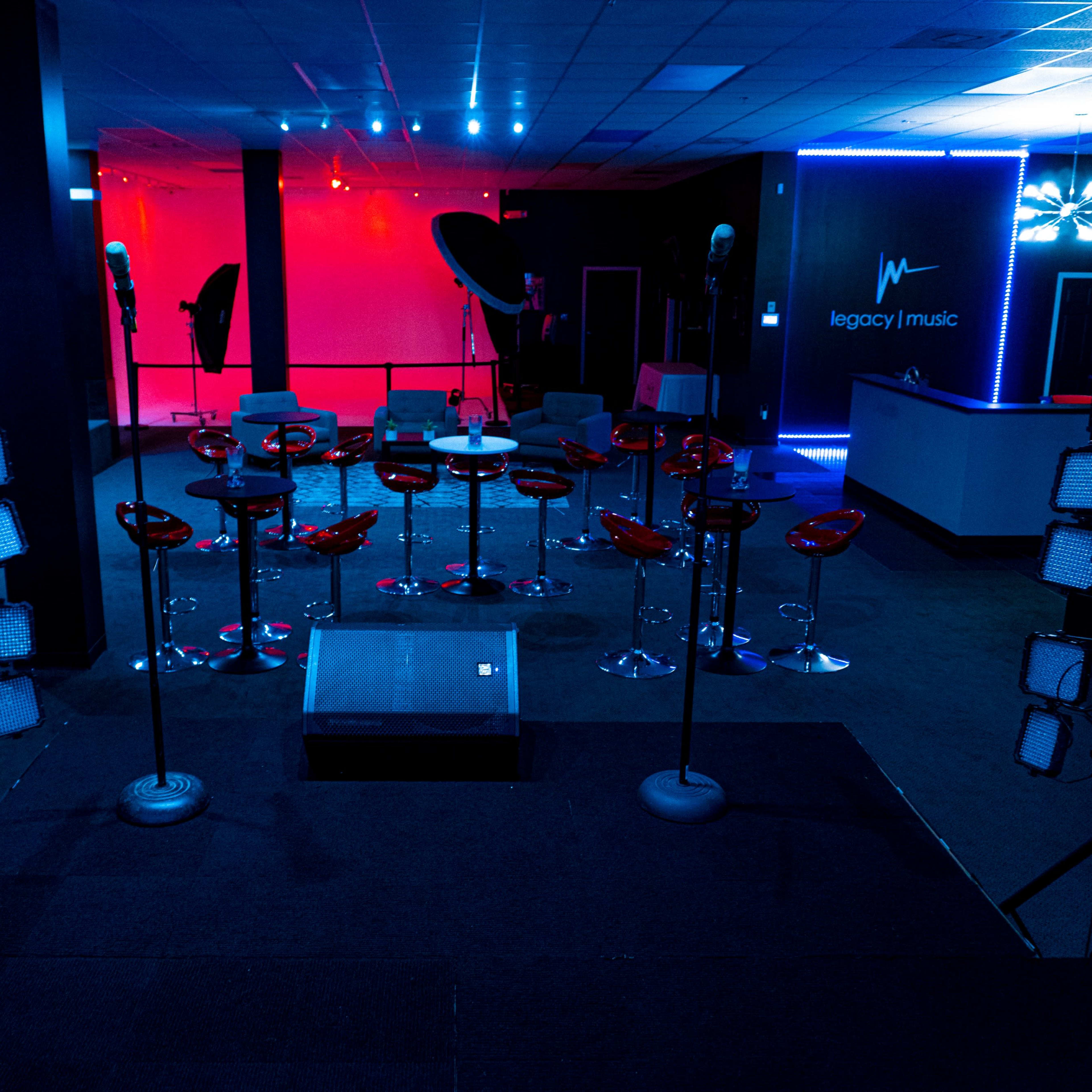 A dimly lit music venue featuring a stage with a sound system, illuminated red and blue lights, and a row of red stools facing the stage.