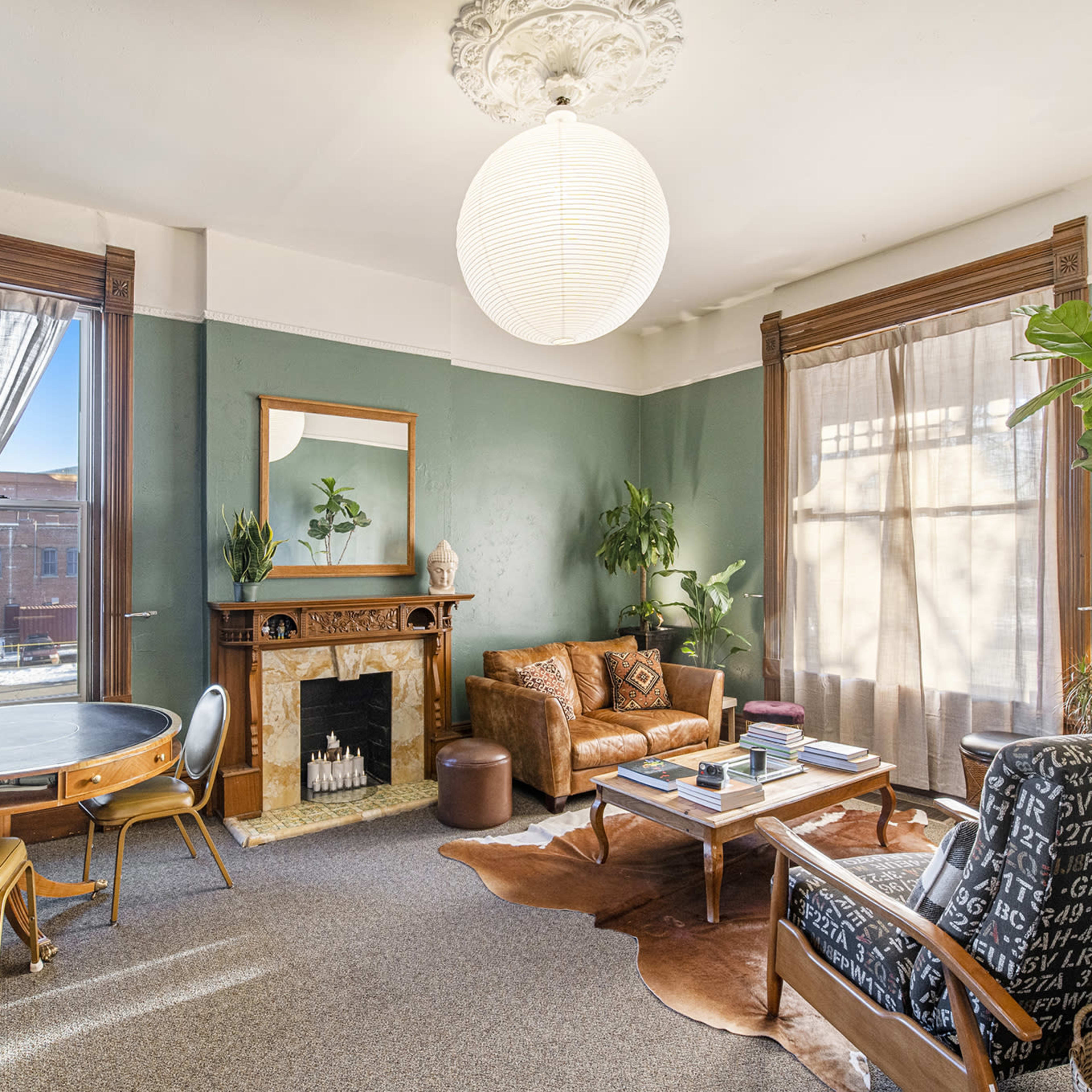 The room features a round table, a leather sofa, and an armchair, with large windows allowing light to illuminate the green walls and a decorative fireplace.