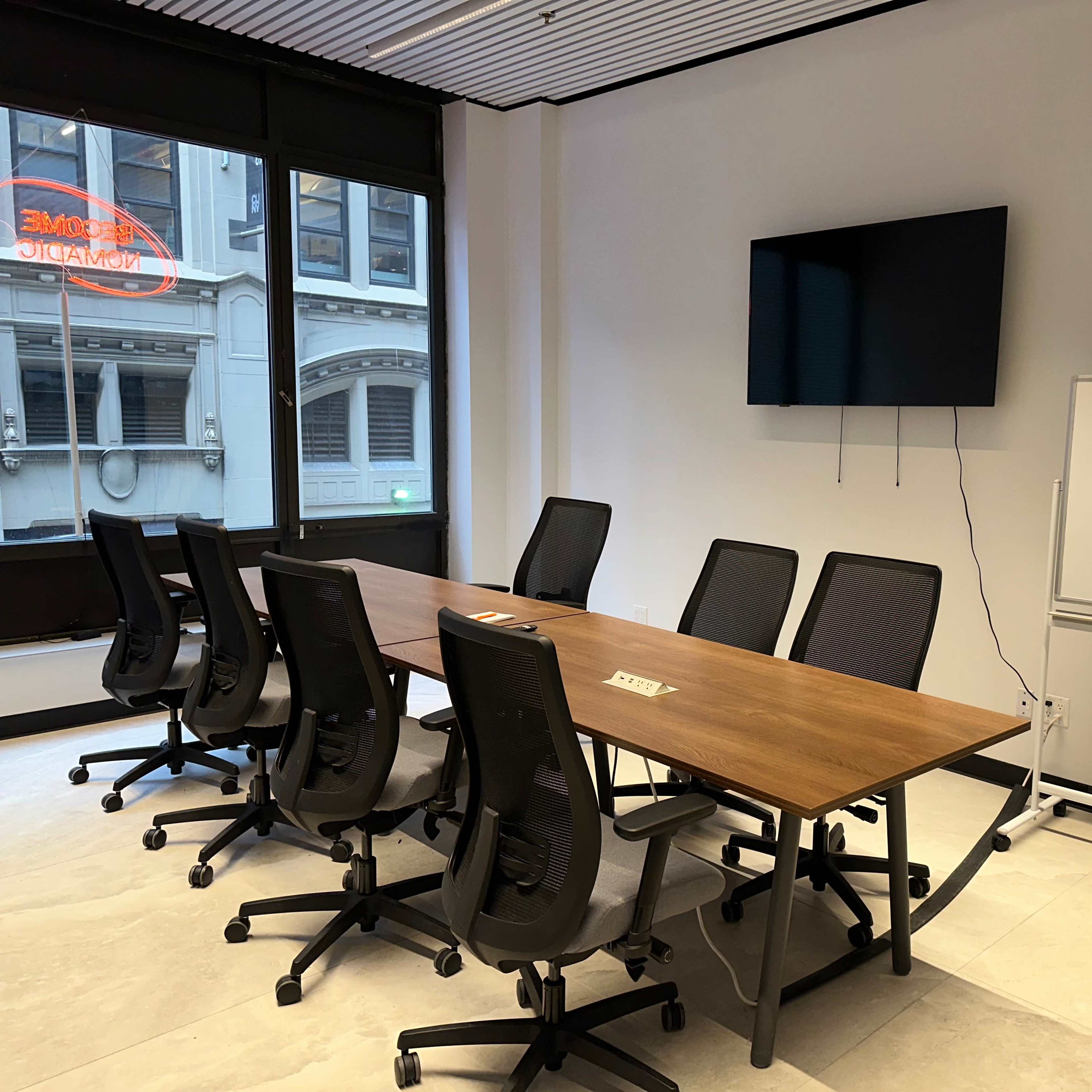 A conference room features a long wooden table surrounded by black ergonomic chairs, a large screen mounted on the wall, and a whiteboard nearby.
