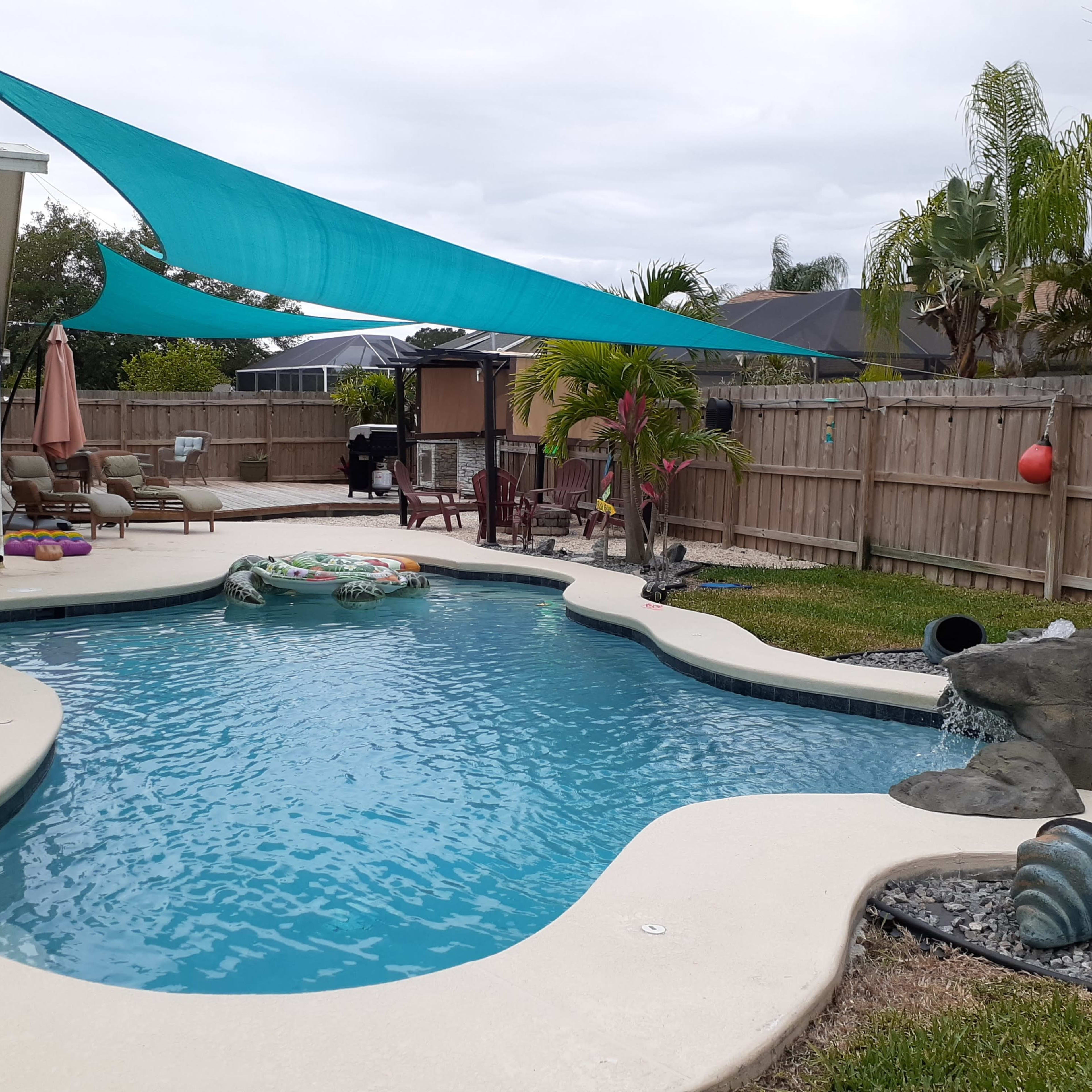 A backyard with a swimming pool surrounded by landscaped grass and palm trees, with a shaded area provided by a blue sail.