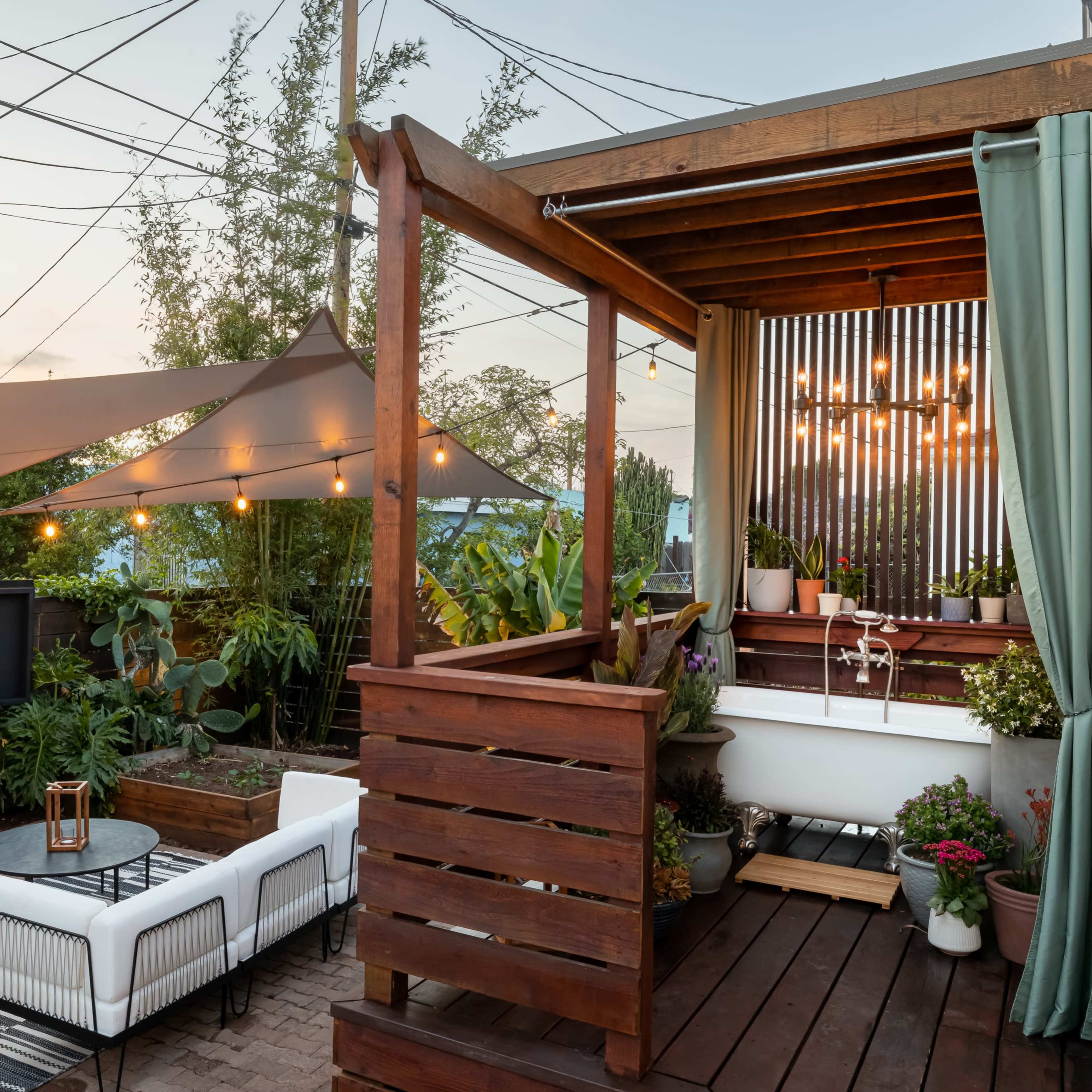 A backyard with a wooden deck, outdoor seating, and a large television, surrounded by plants and illuminated by string lights.