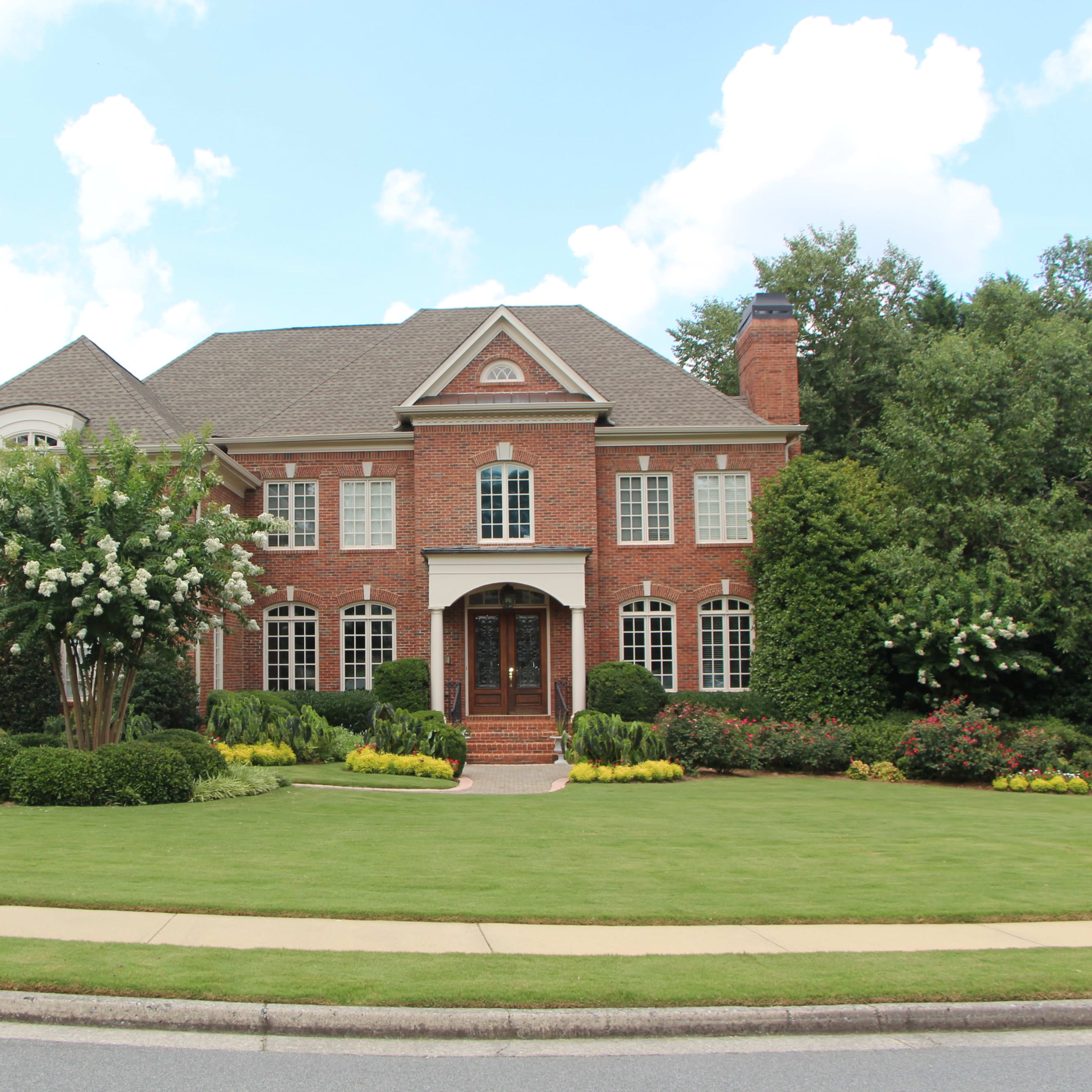A large, red-brick two-story house surrounded by neatly trimmed hedges and a well-maintained lawn.