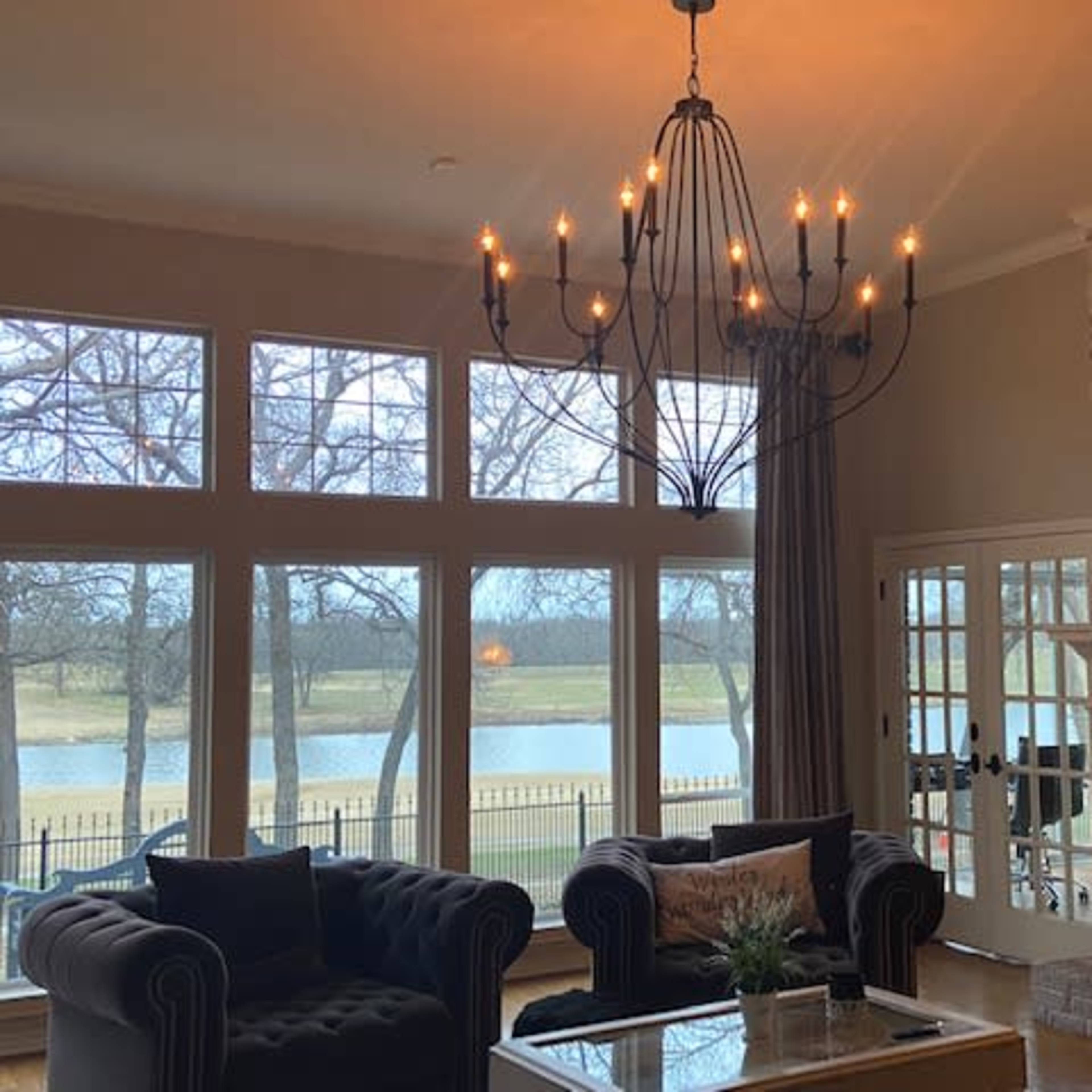 A living room features large windows overlooking a lake, with a chandelier hanging above a coffee table and two tufted sofas.