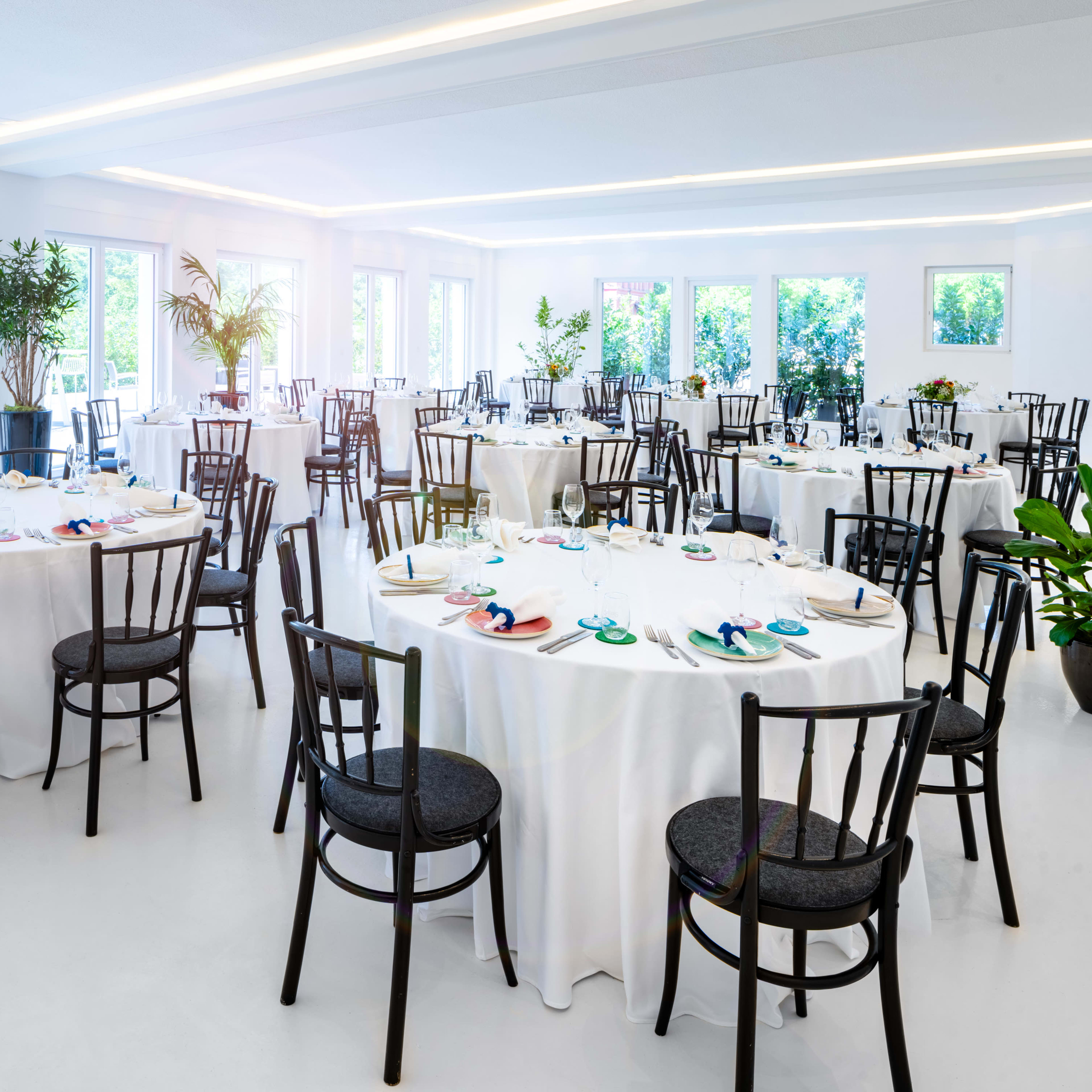 A spacious dining room features several round tables set with white tablecloths and black chairs, surrounded by greenery and large windows.