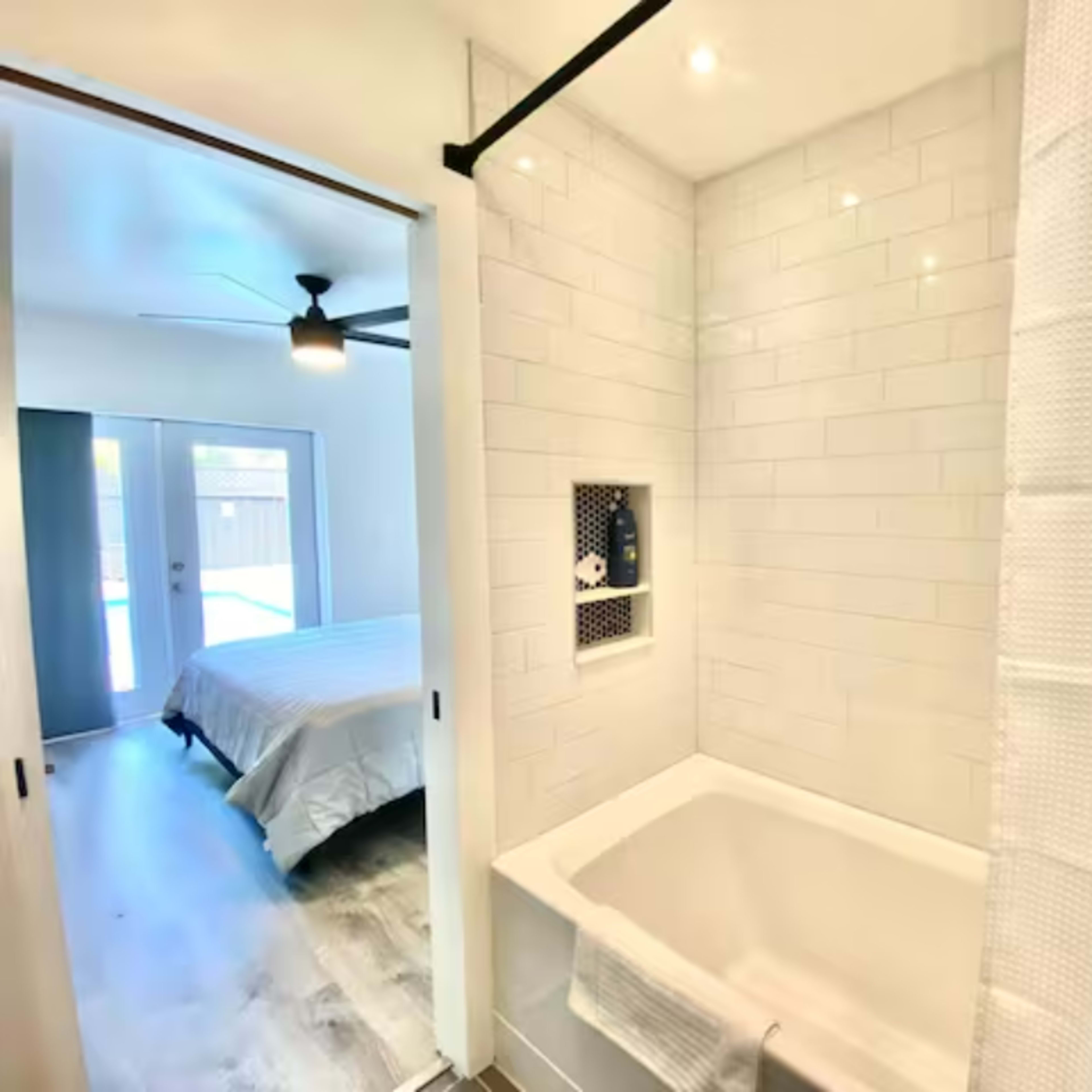 A bright bathroom features a white tiled shower area with a bath tub and an open doorway leading to a bedroom.