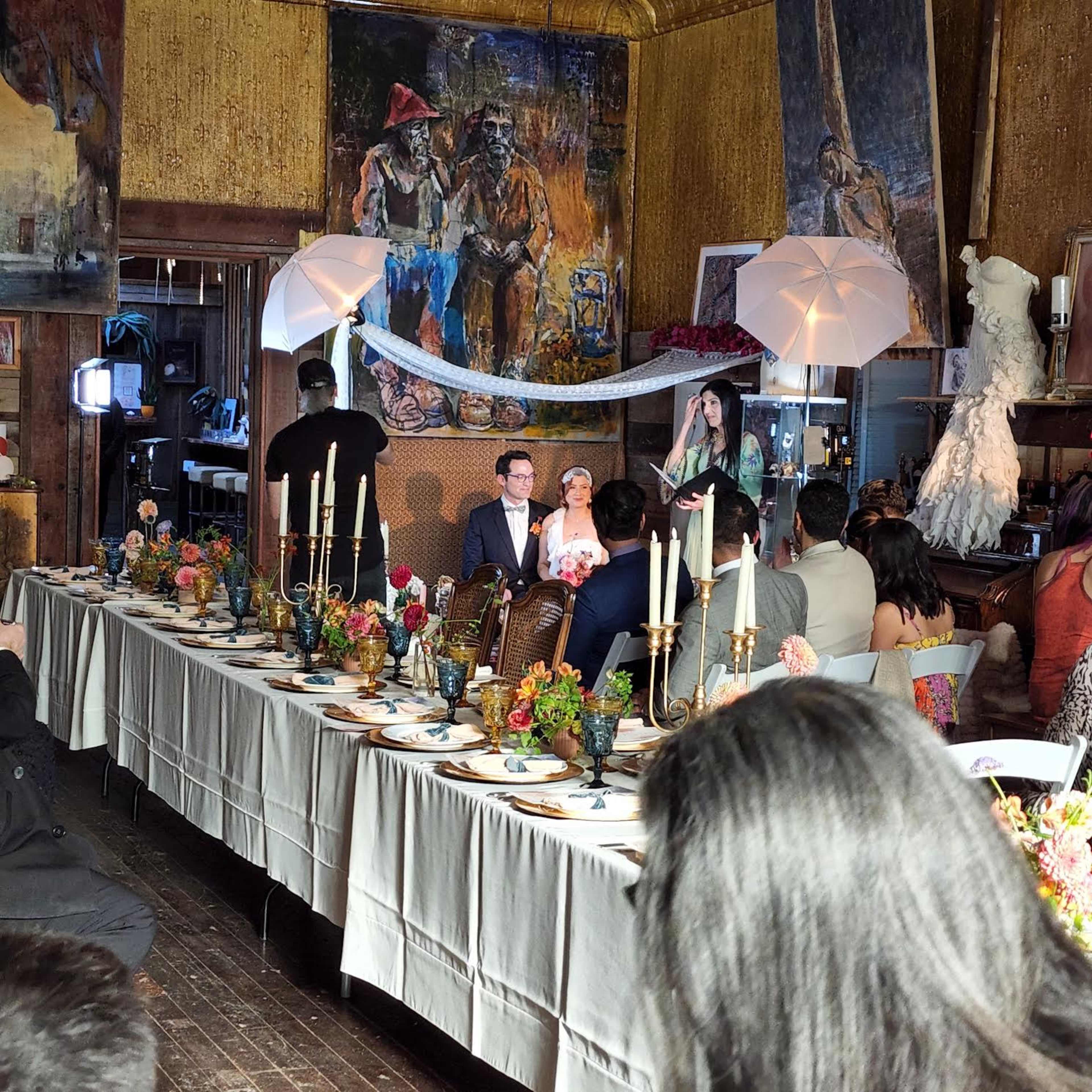A couple sits at the head of a long dining table adorned with candles and flowers, while guests gather around in a decorated room featuring artwork on the walls.
