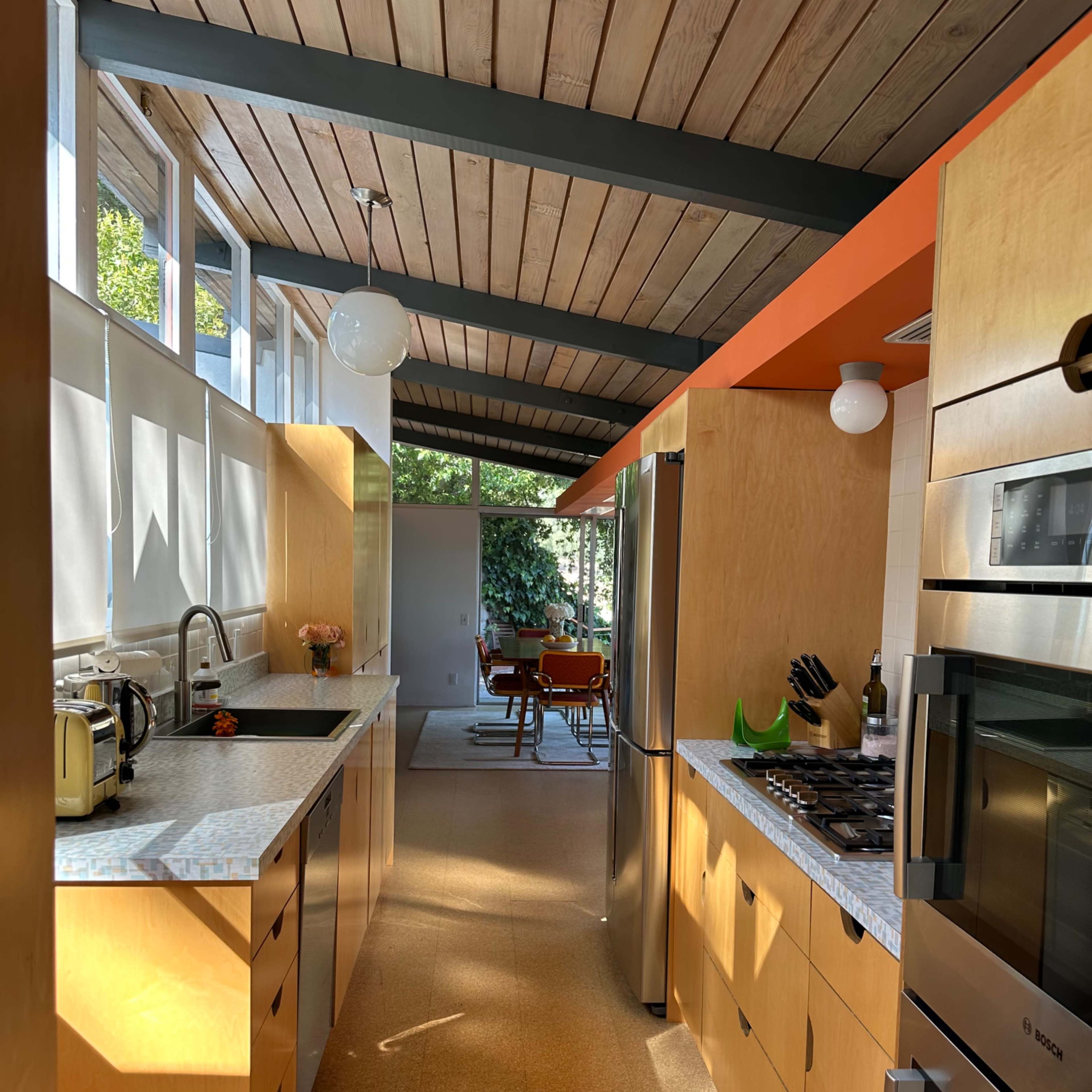A modern kitchen with wooden cabinetry, a marble countertop, and stainless steel appliances, leading into a dining area with a view of greenery.