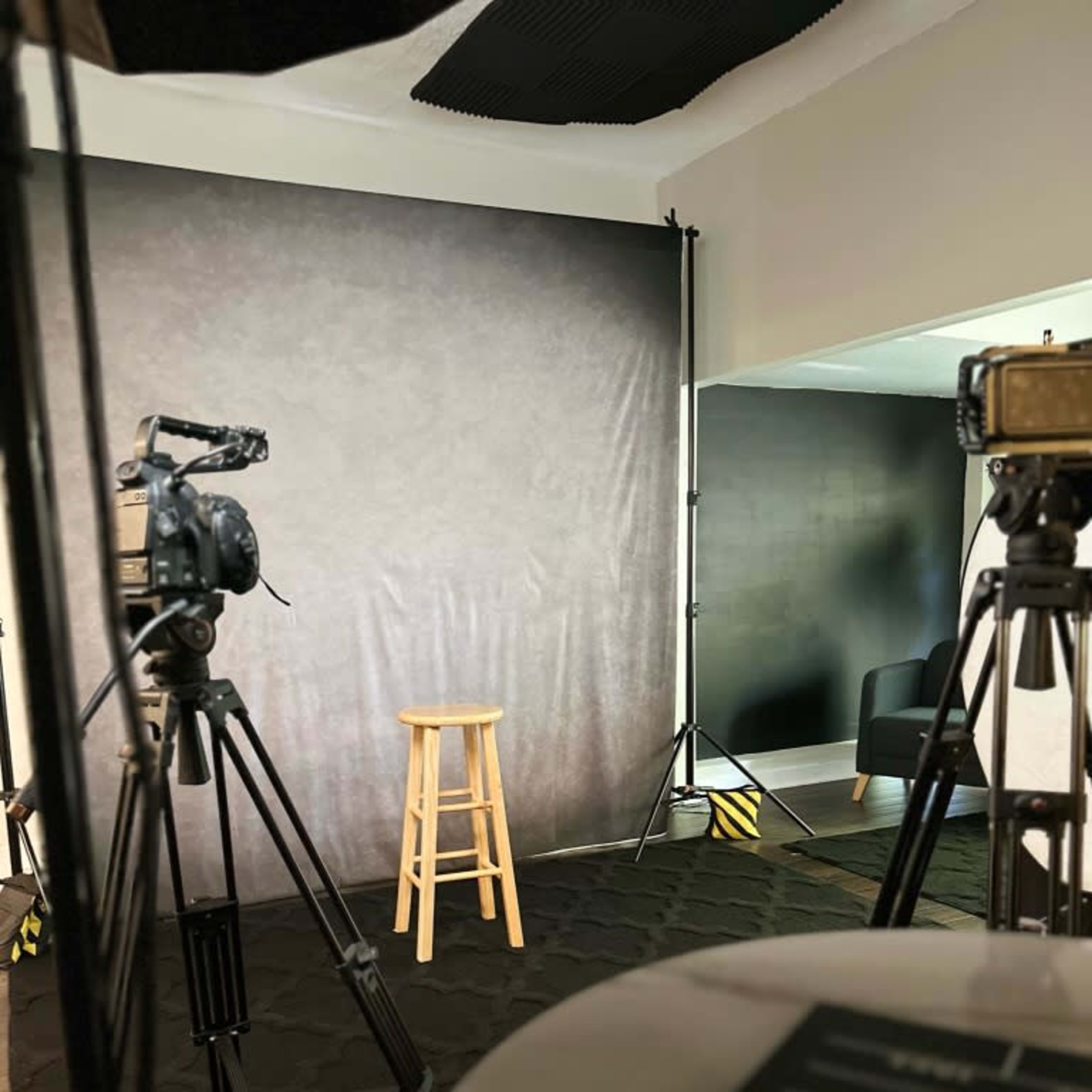 A photography or video recording setup featuring a stool in front of a dark backdrop, with several cameras and tripods positioned around the scene.