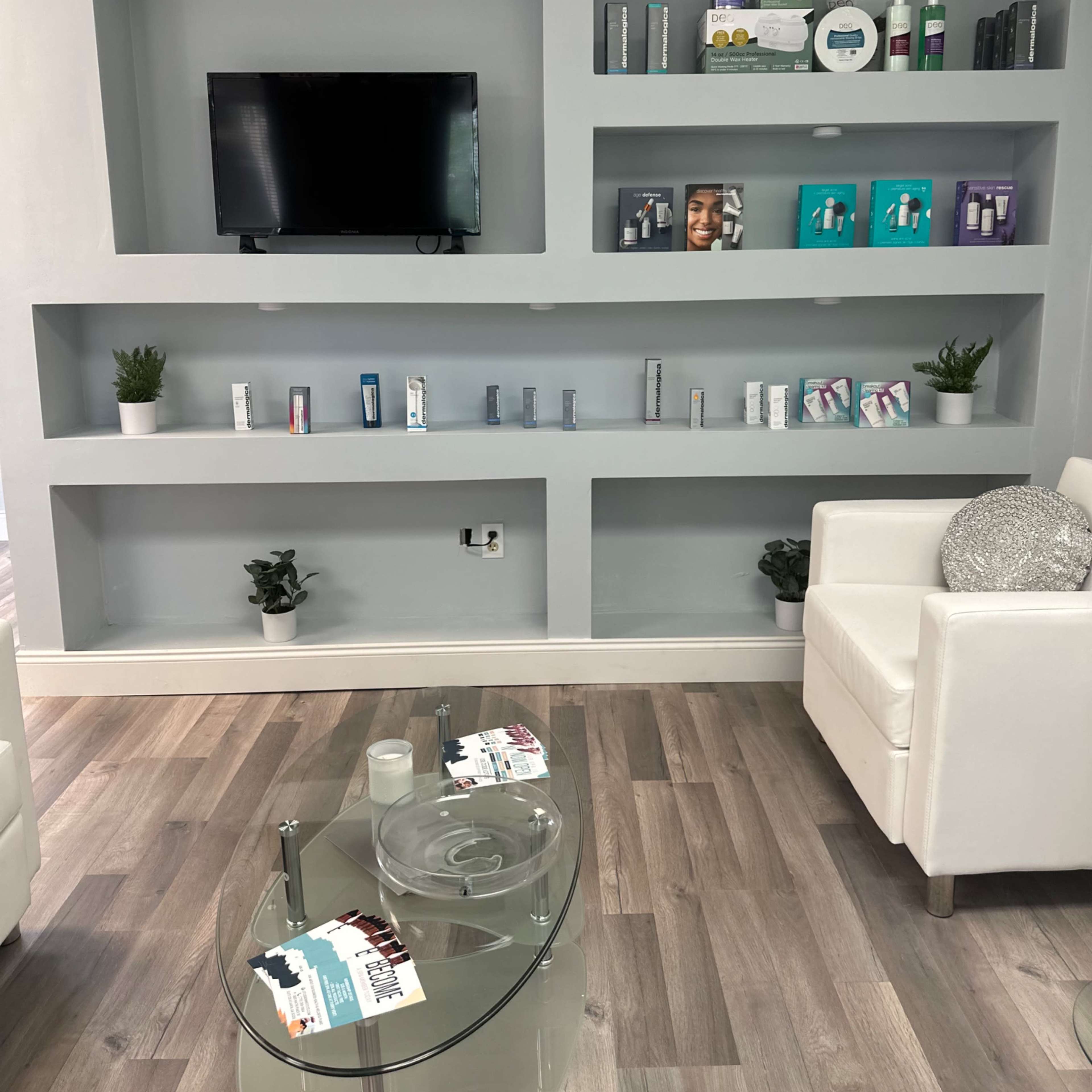 A modern waiting area with a wall-mounted television, neatly arranged shelves displaying products, and white furniture surrounding a glass coffee table.