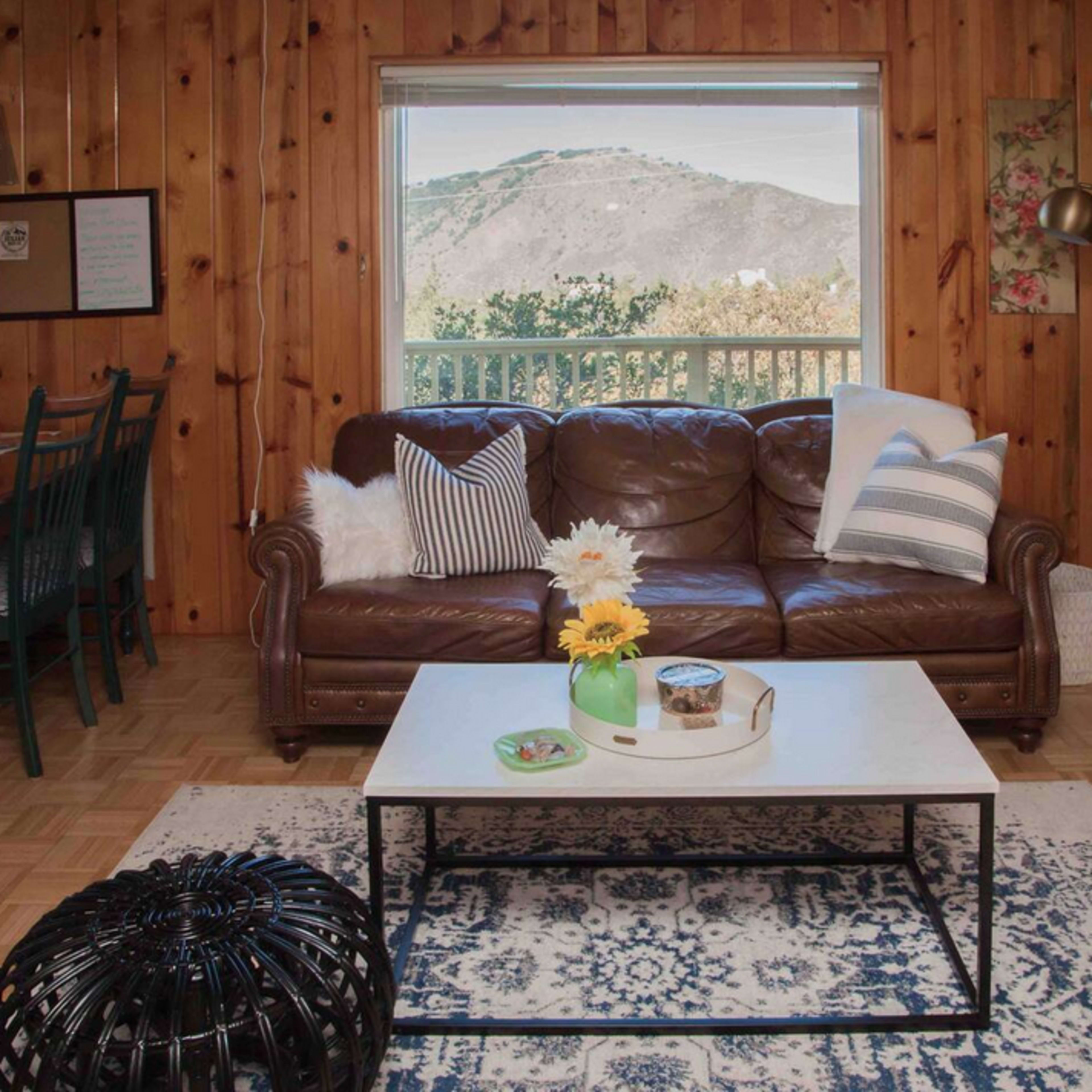 A cozy living room with a brown leather sofa, a coffee table, and a large window displaying a mountain view.