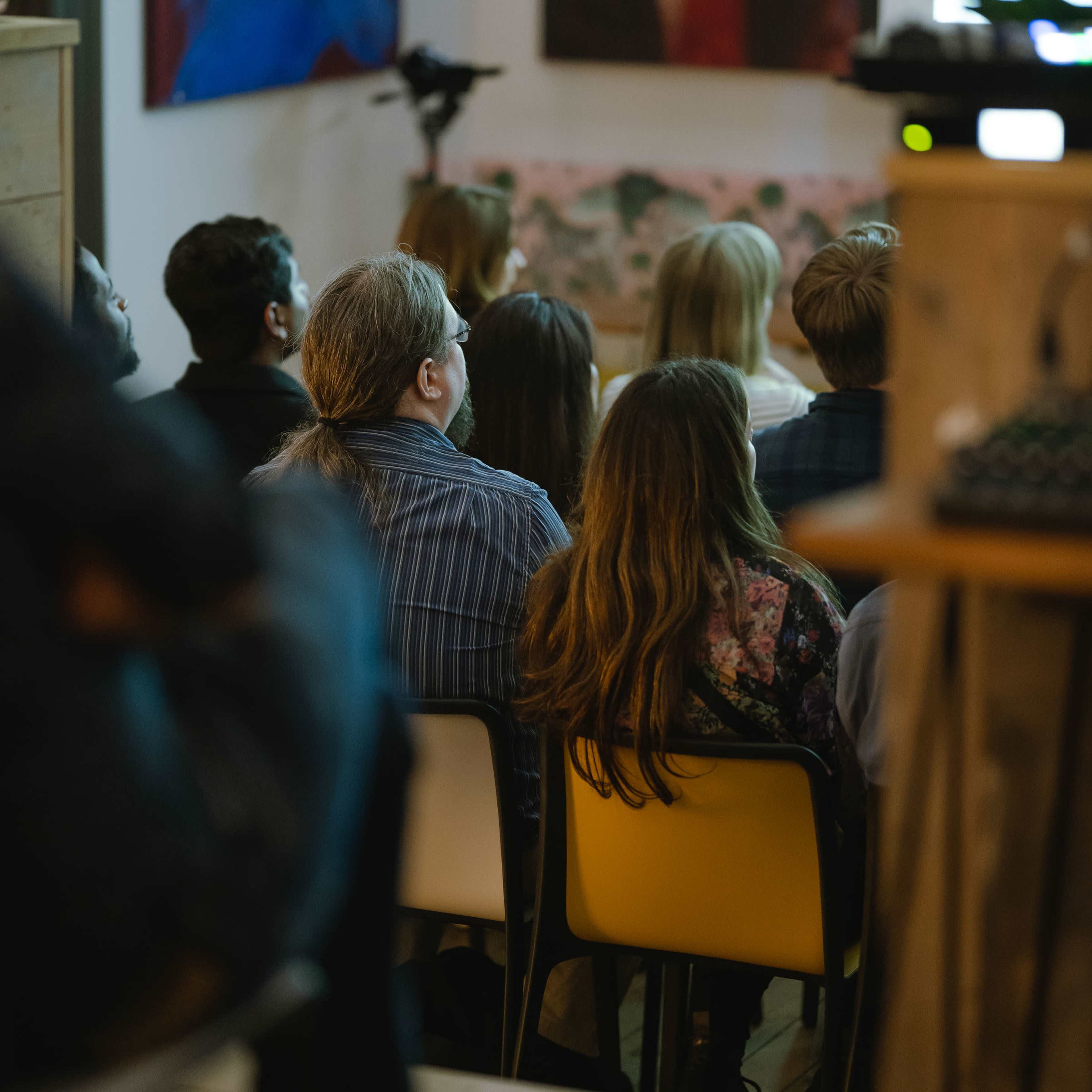 A group of people is seated in a dimly lit room, facing a presentation screen while various abstract paintings decorate the walls.