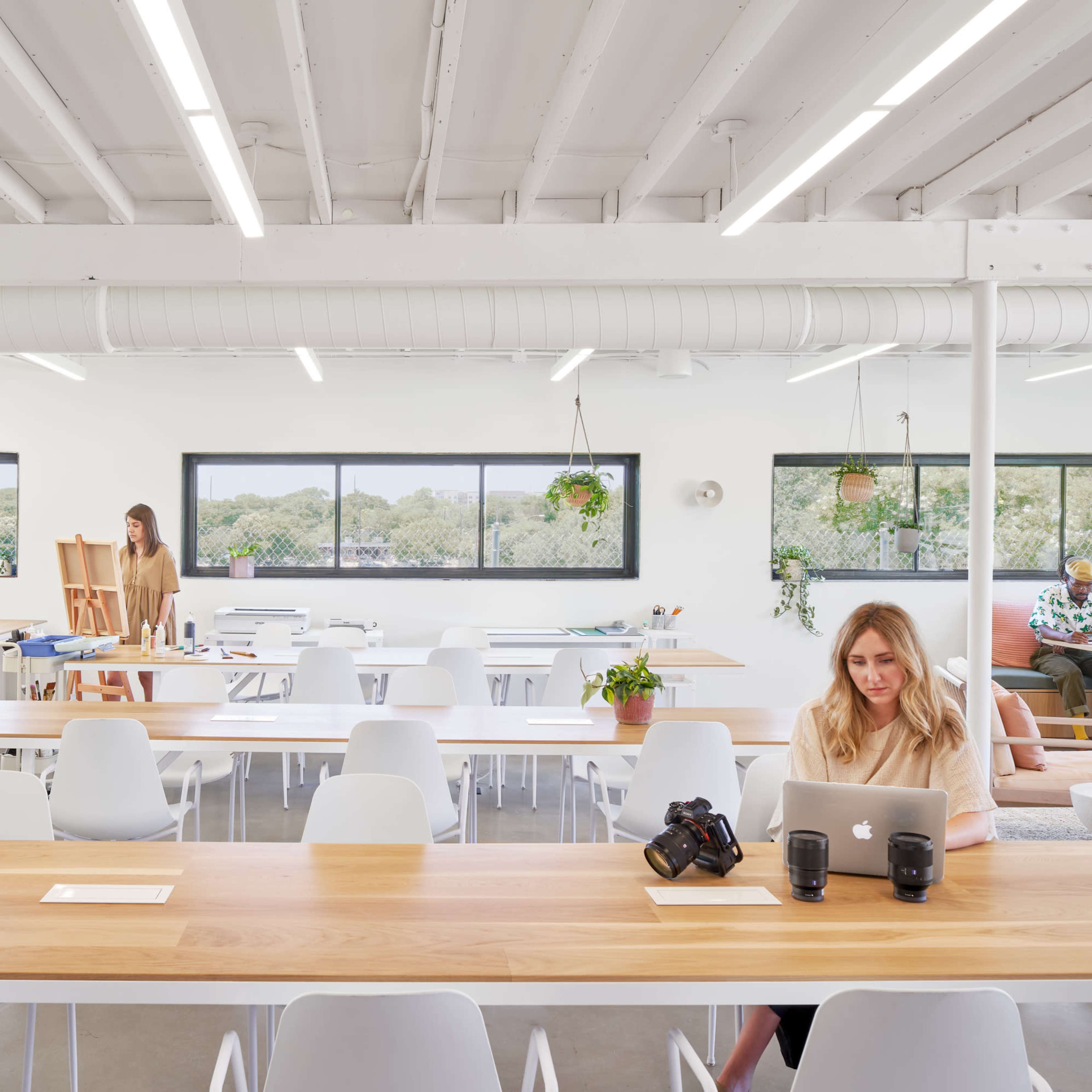 A bright, modern workspace features several tables, people engaged in different activities, and large windows allowing natural light to fill the room.