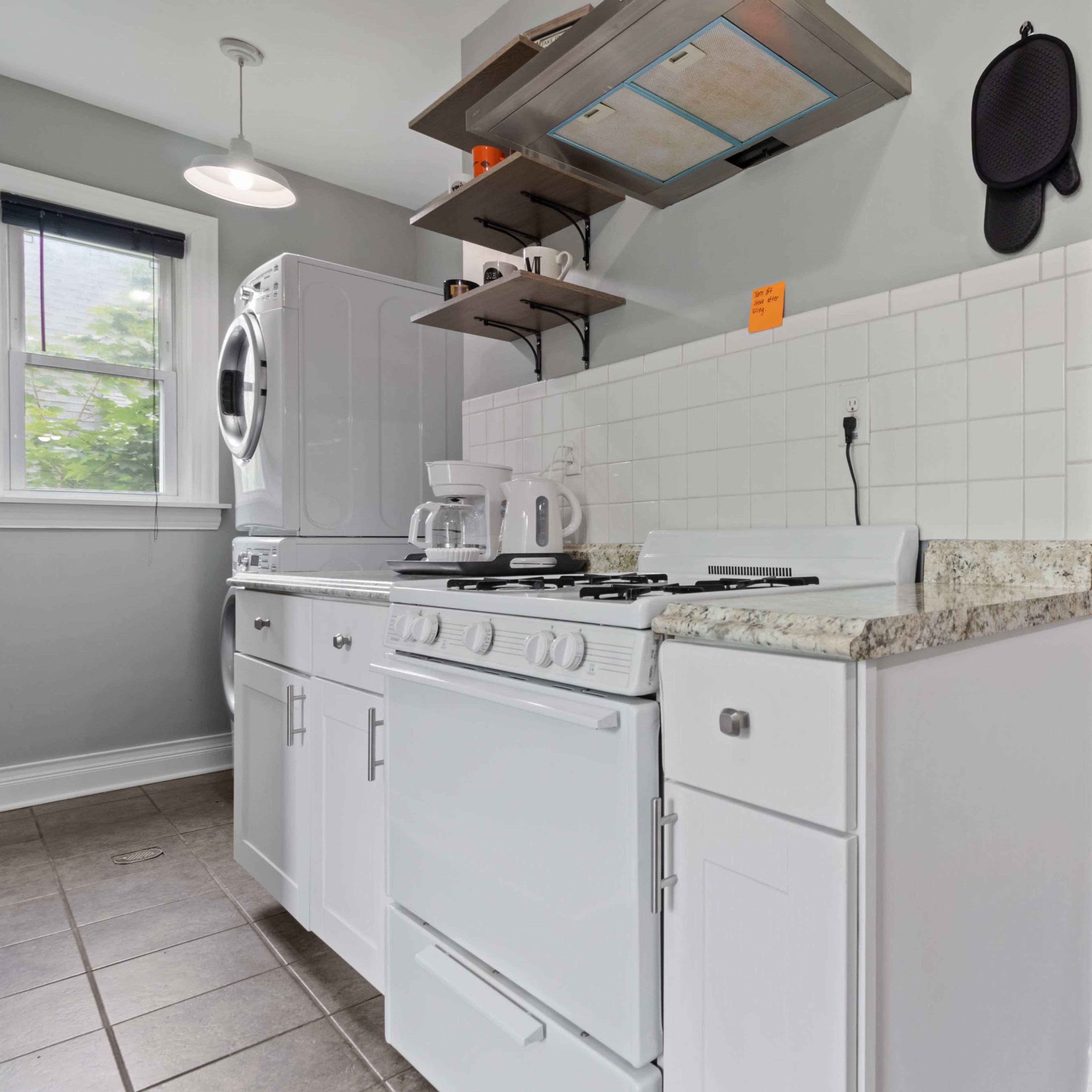 A compact kitchen with a white gas stove, a wall-mounted shelf holding kitchen items, and a washer dryer unit beside it.
