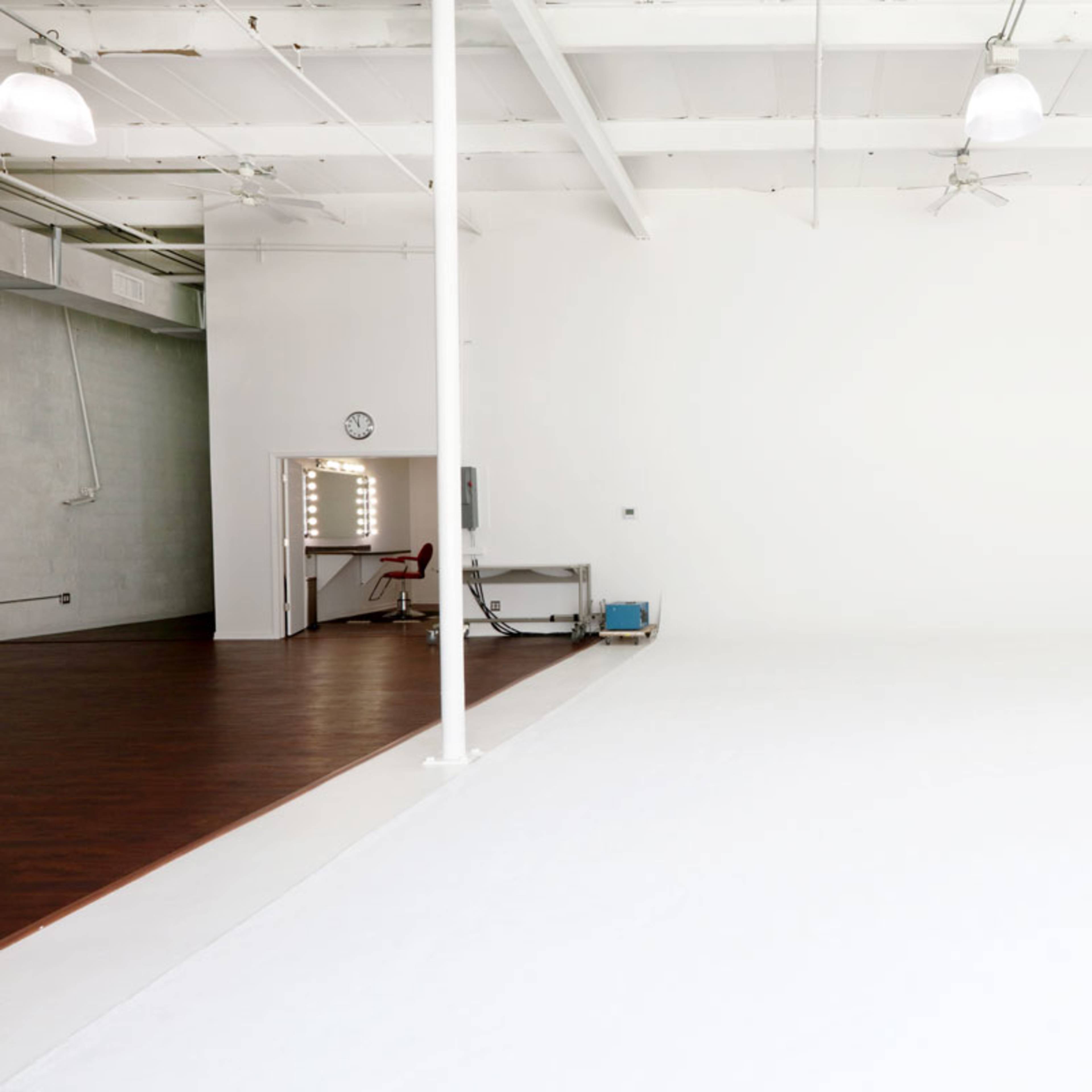 A bright, empty studio space with white walls and a wooden floor, featuring a small red chair and a mirror with lights in the background.