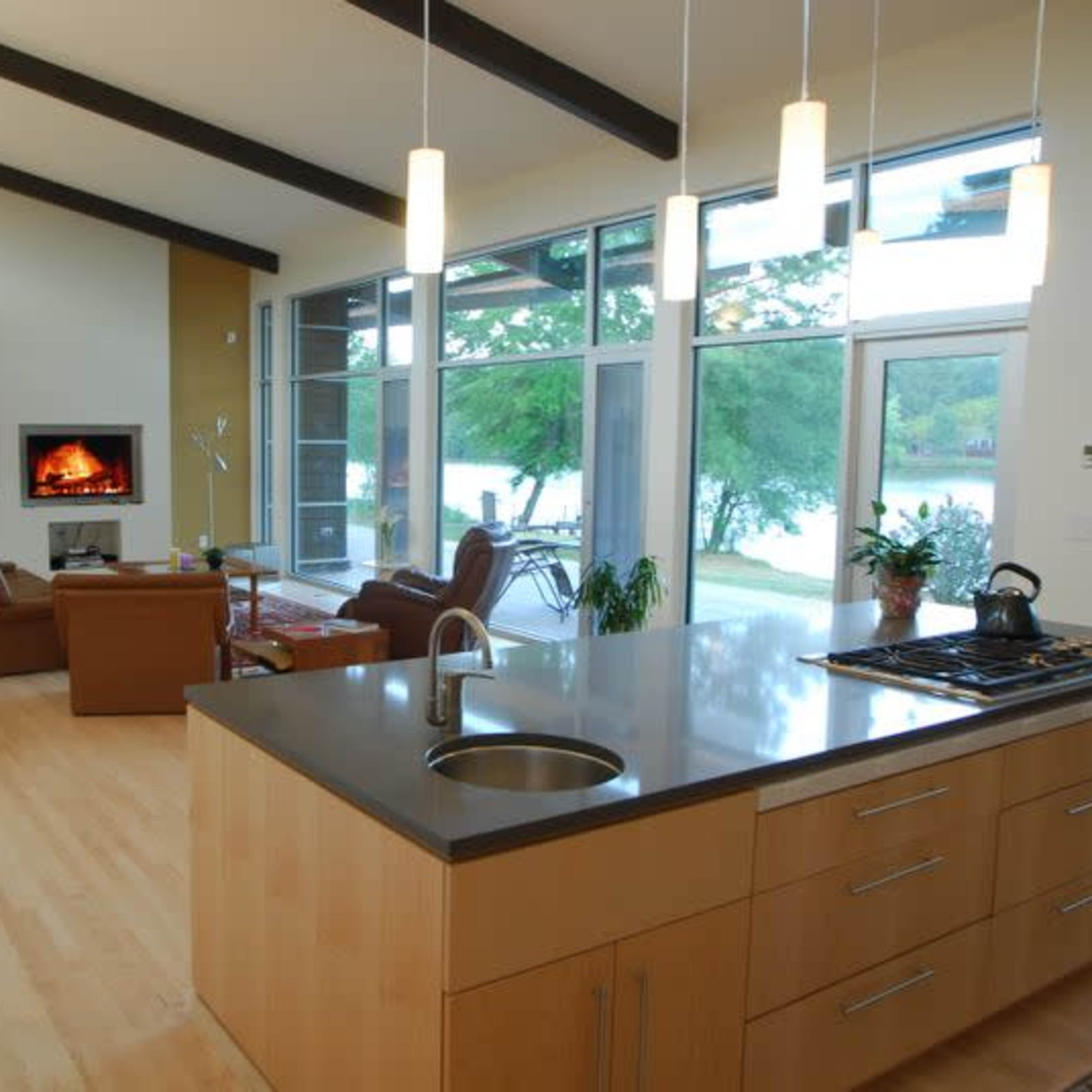 A modern open-concept kitchen and living area with large windows overlooking a lake, featuring a central island and a fireplace in the living space.