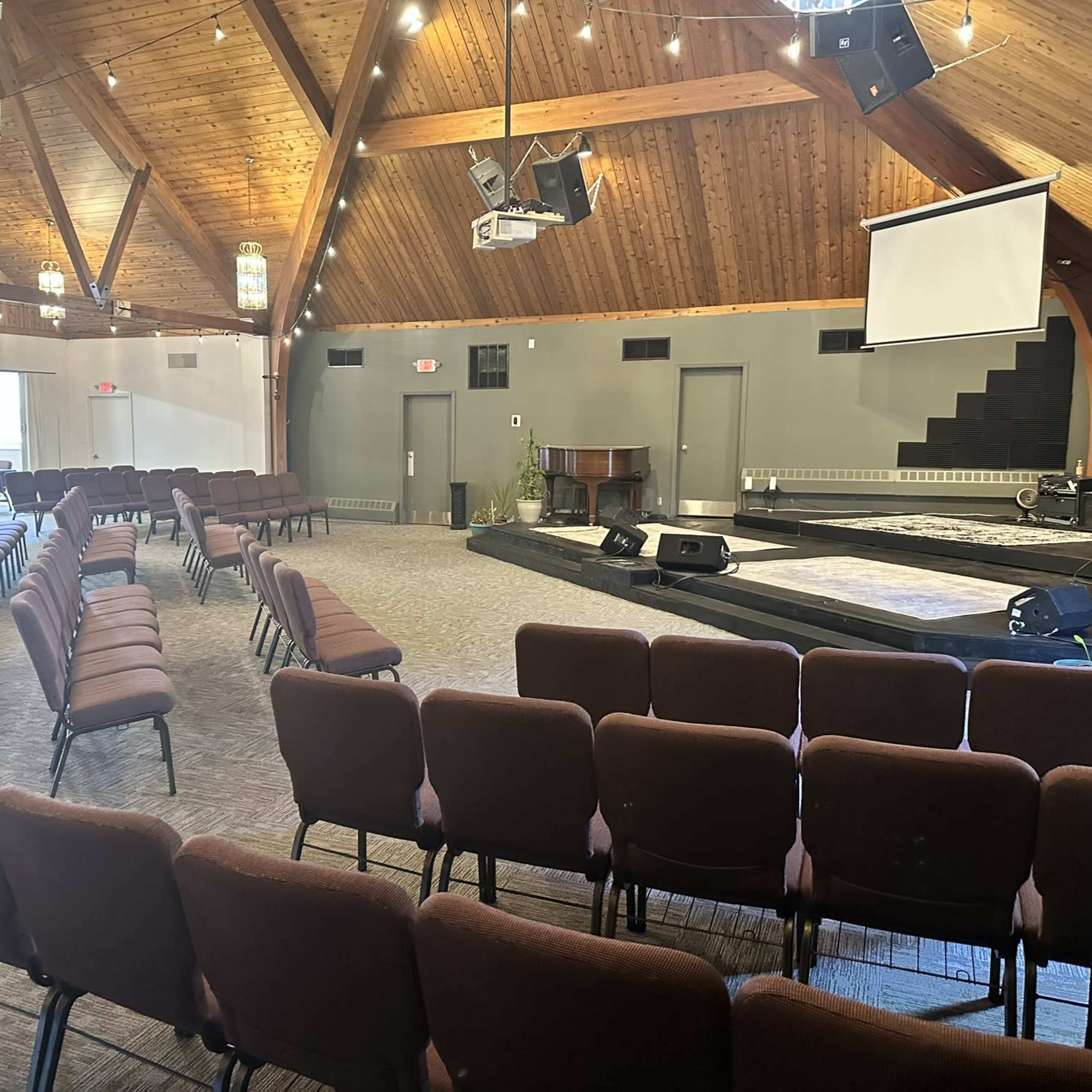 An empty indoor auditorium with wooden beams, rows of brown chairs facing a stage, and a large screen on the wall.