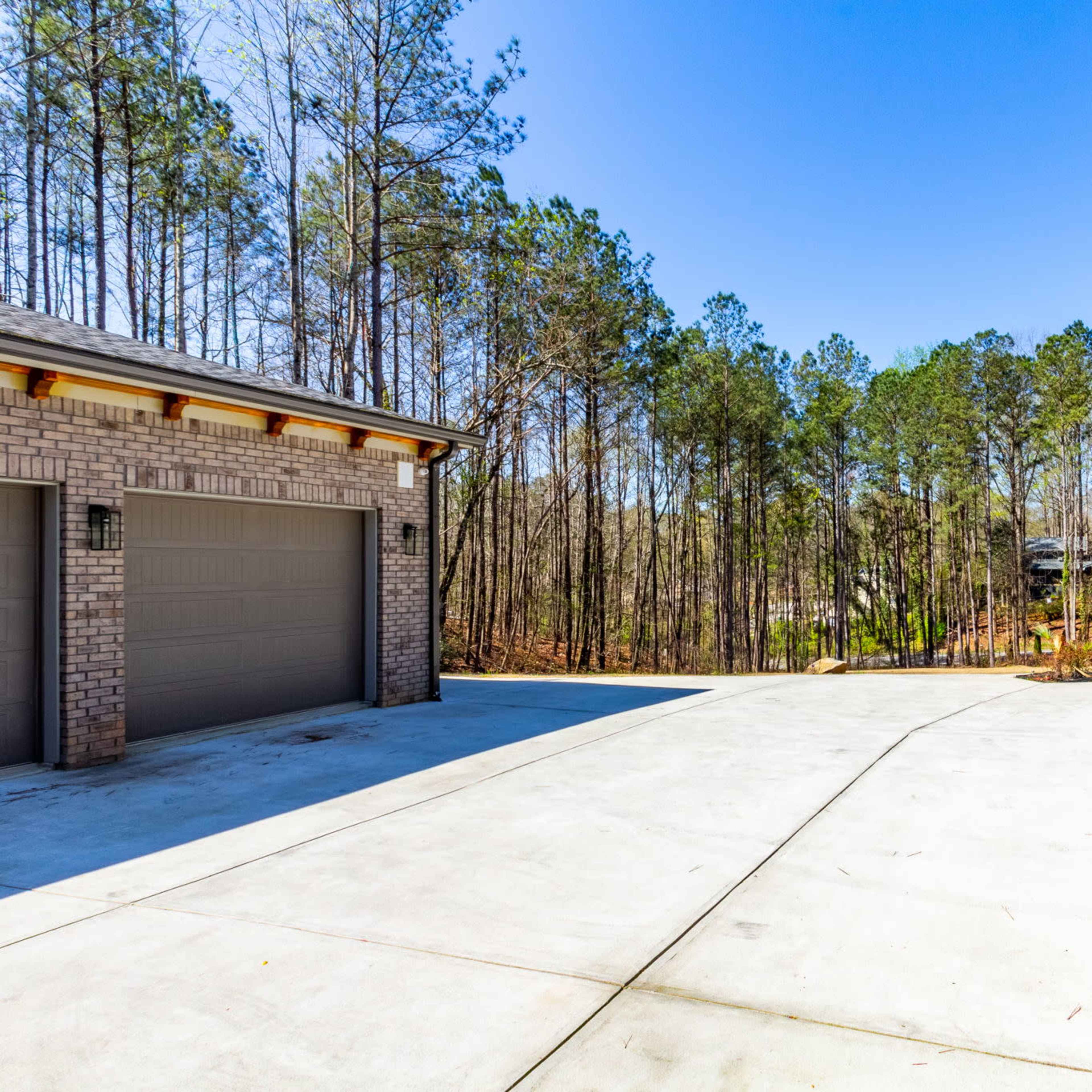 A concrete driveway leads to a house surrounded by tall pine trees under a clear blue sky.