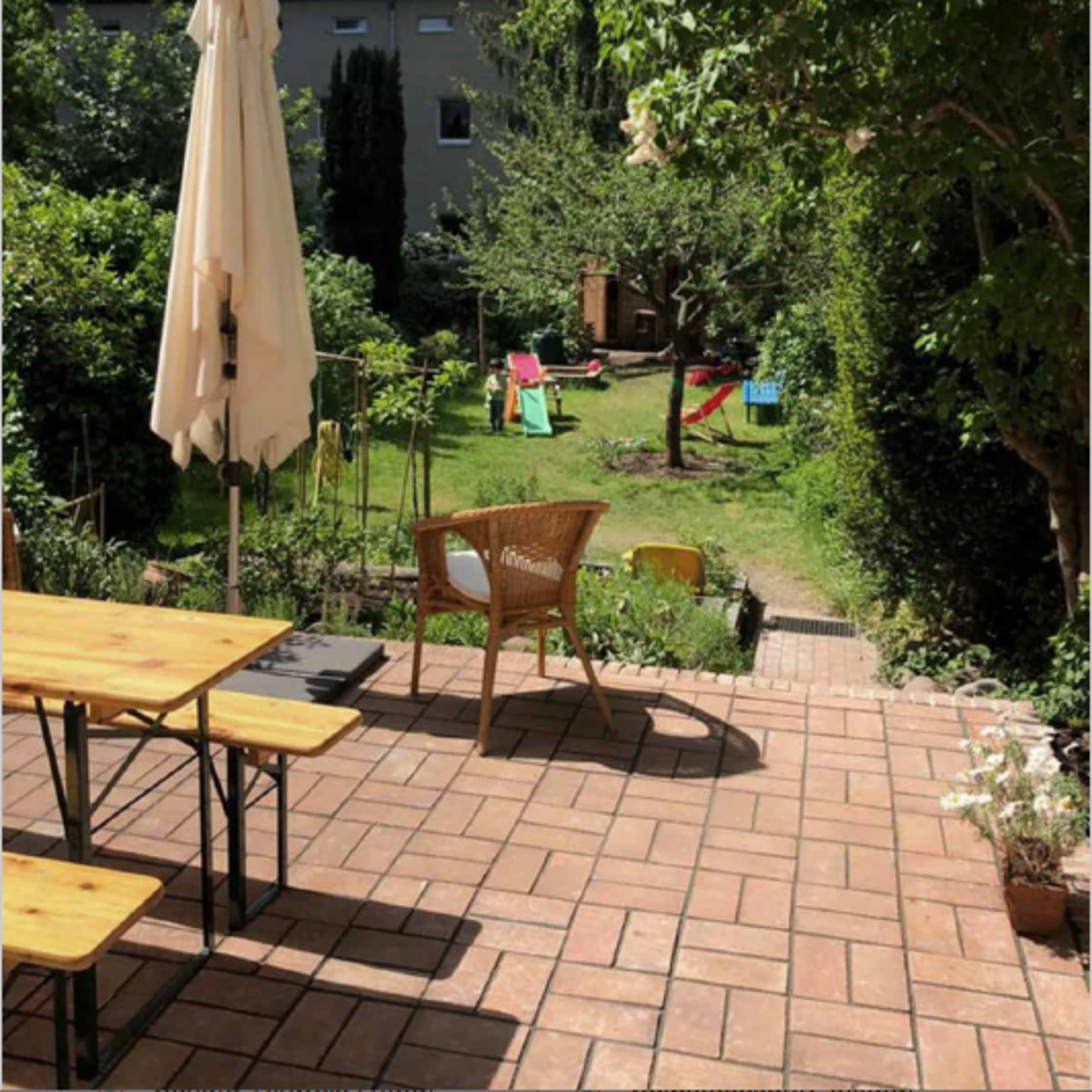 A sunlit patio with a table and chairs overlooks a green garden featuring lounge chairs and greenery.