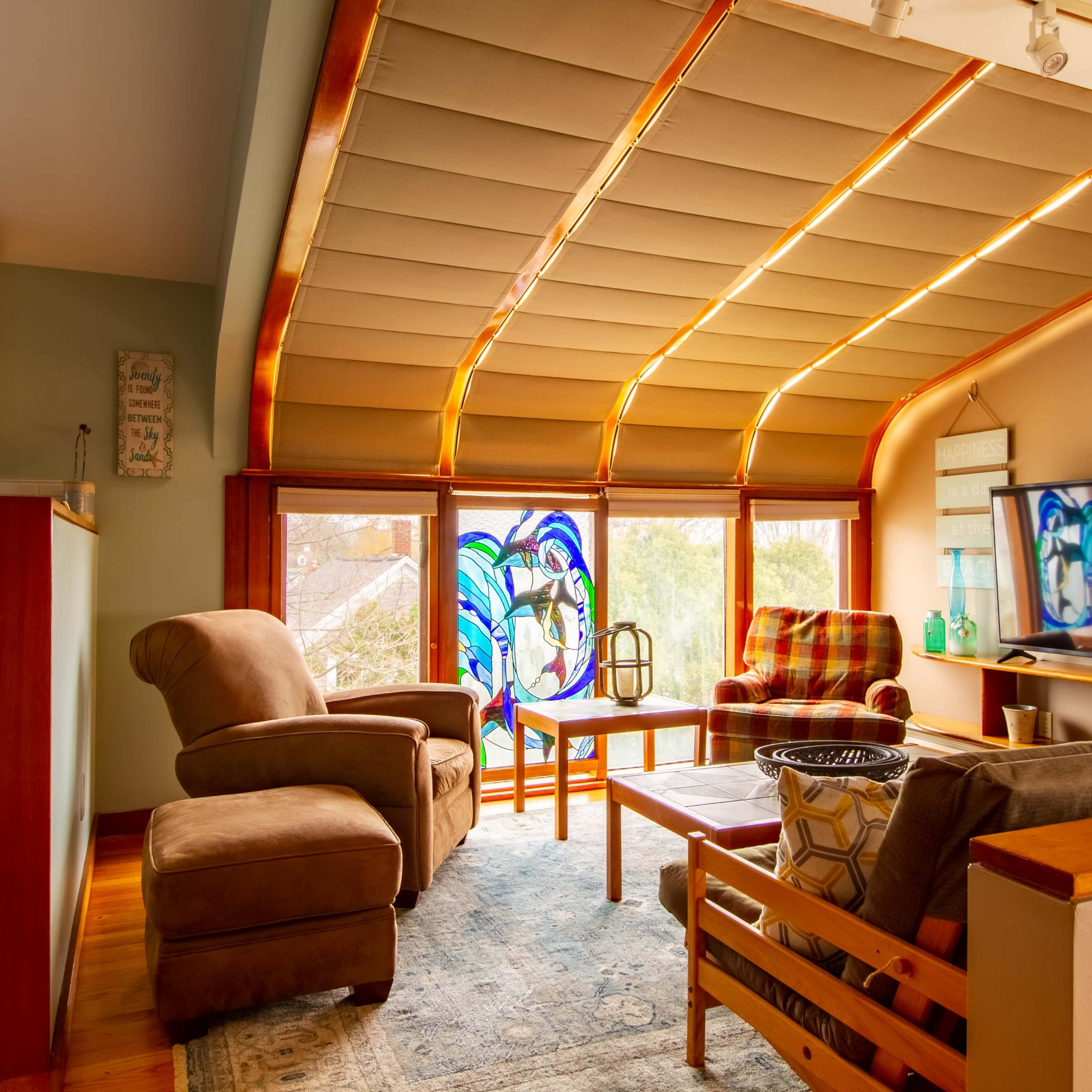 The scene features a cozy living area with a combination of seating, a large window adorned with stained glass, and a television mounted on the wall.