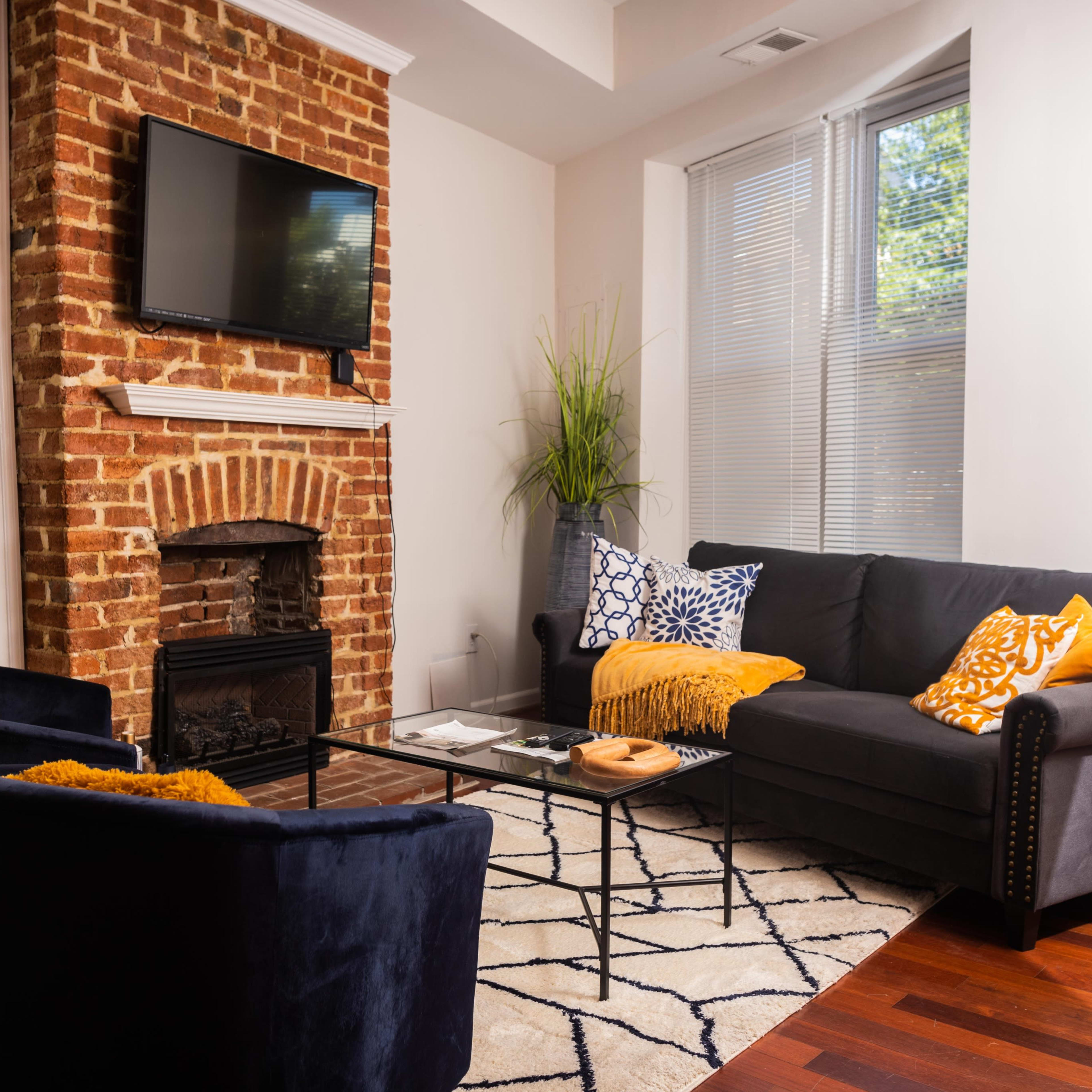 A cozy living room features a brick fireplace, a wall-mounted television, a dark gray sofa, and two blue armchairs, all arranged around a glass coffee table on a patterned rug.
