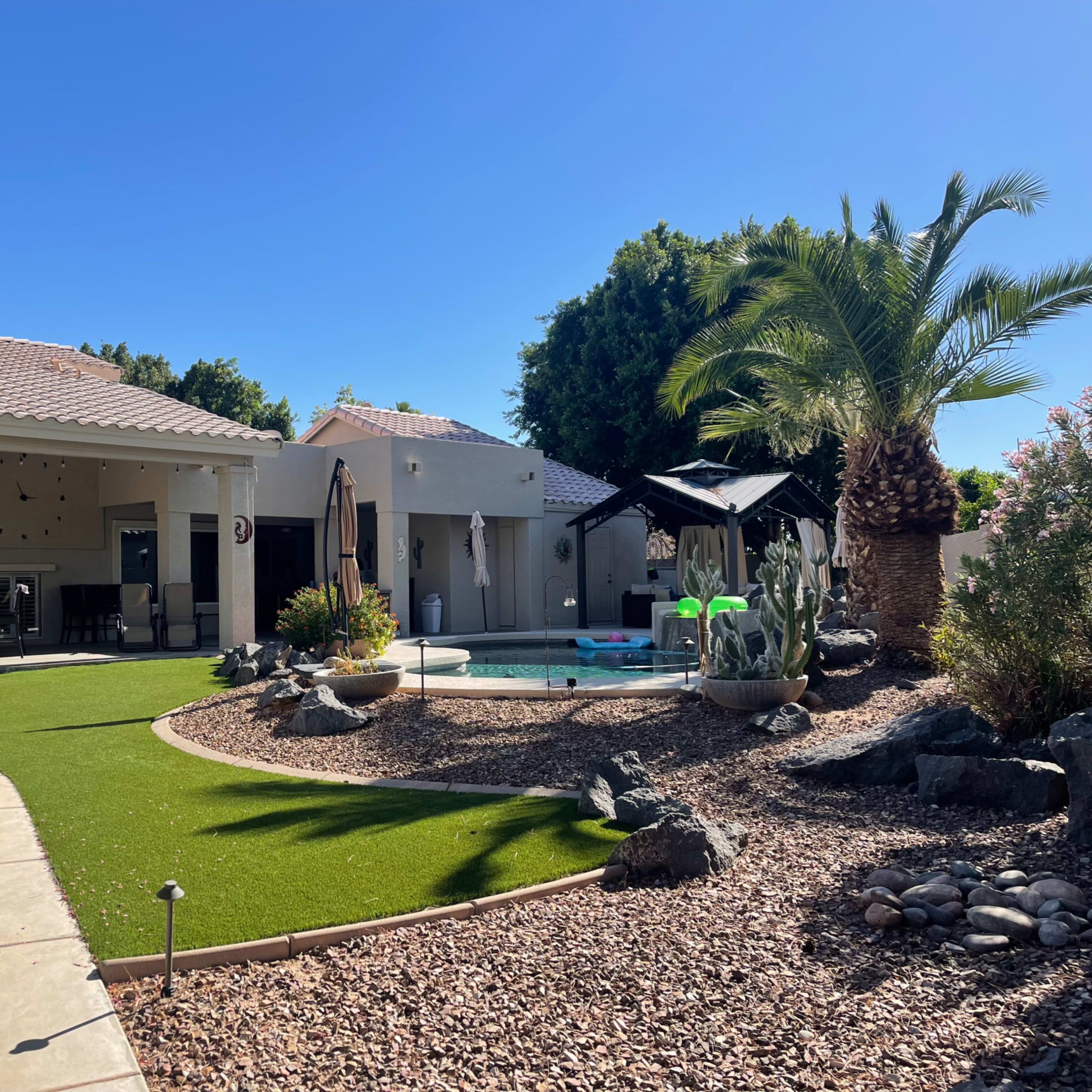 A landscaped backyard with a pool, palm trees, and a gazebo surrounded by gravel and ornamental plants.
