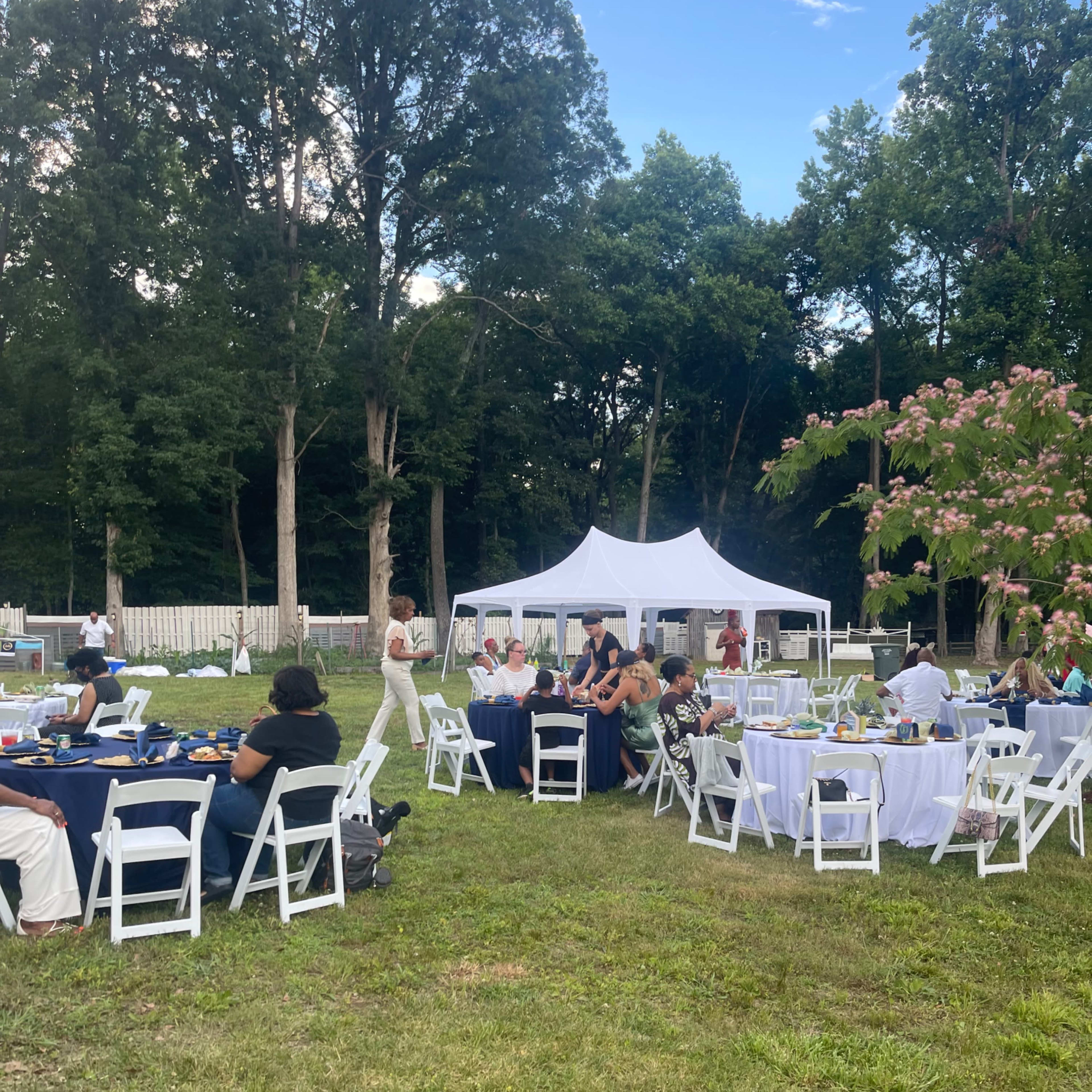 A large outdoor gathering features guests seated at round and rectangular tables beneath a white tent, set against a backdrop of trees.