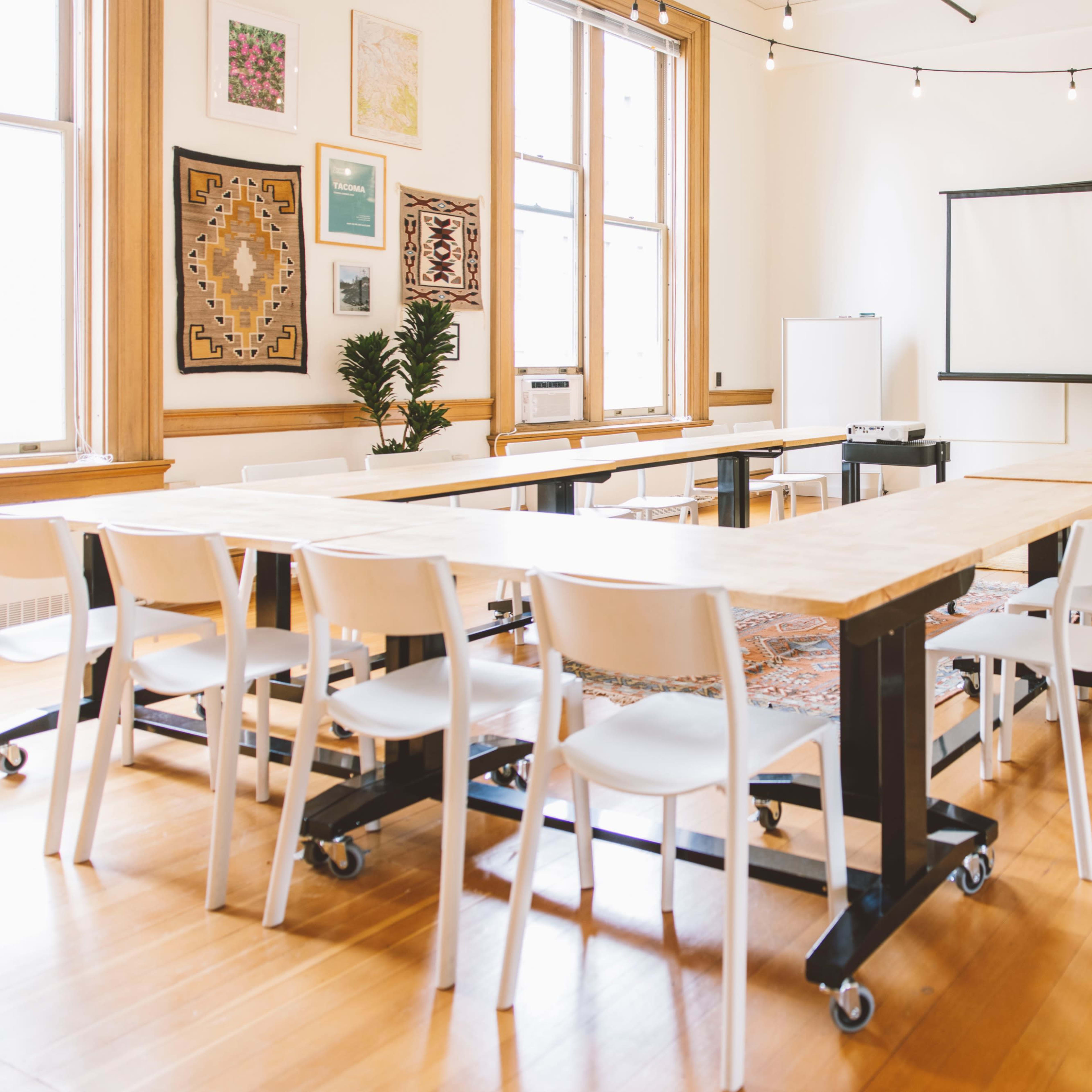 A meeting room features long tables with white chairs, a projector screen, and large windows letting in natural light.