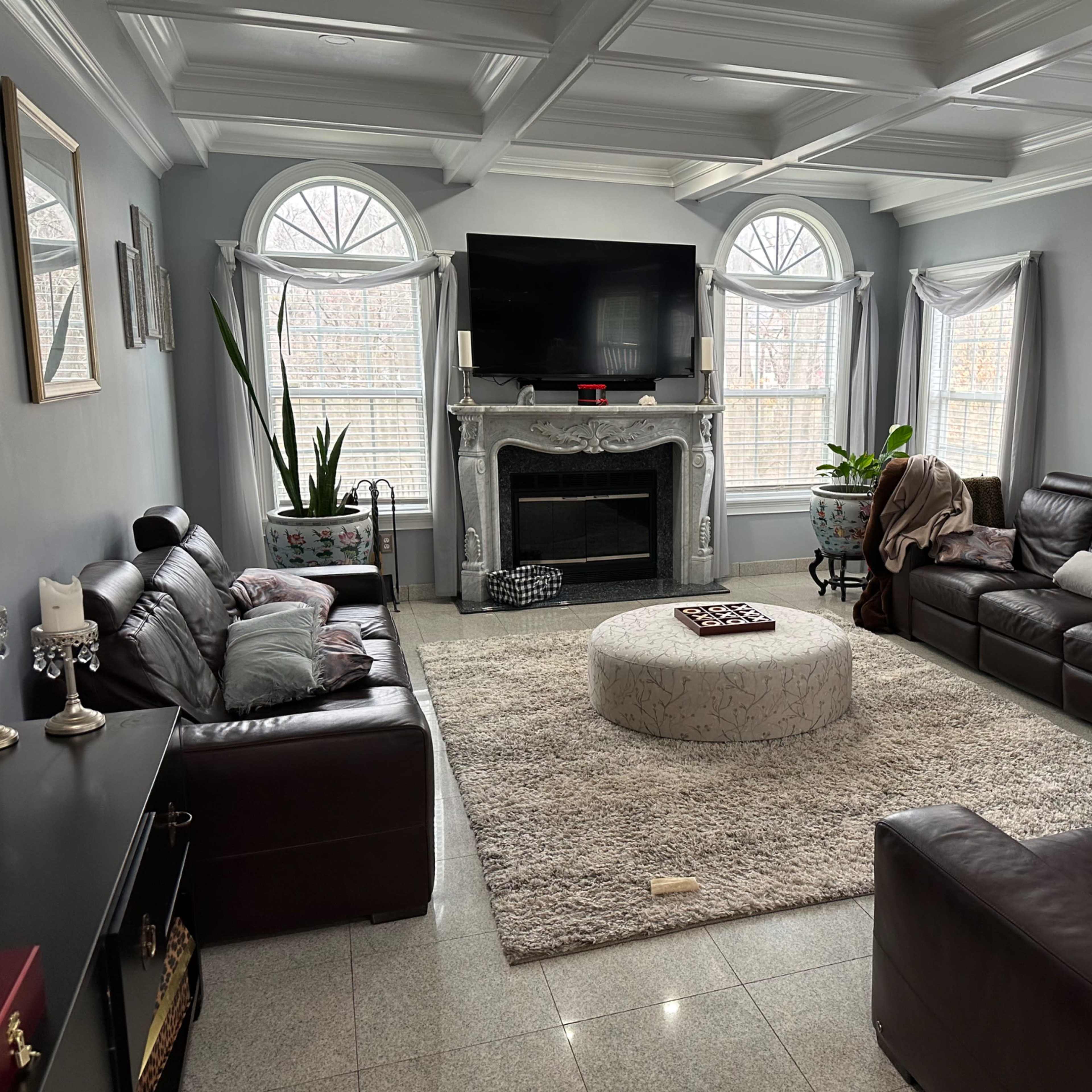 The living room features two dark leather sofas facing a central circular coffee table, with a large television mounted above a decorative fireplace and windows allowing natural light.