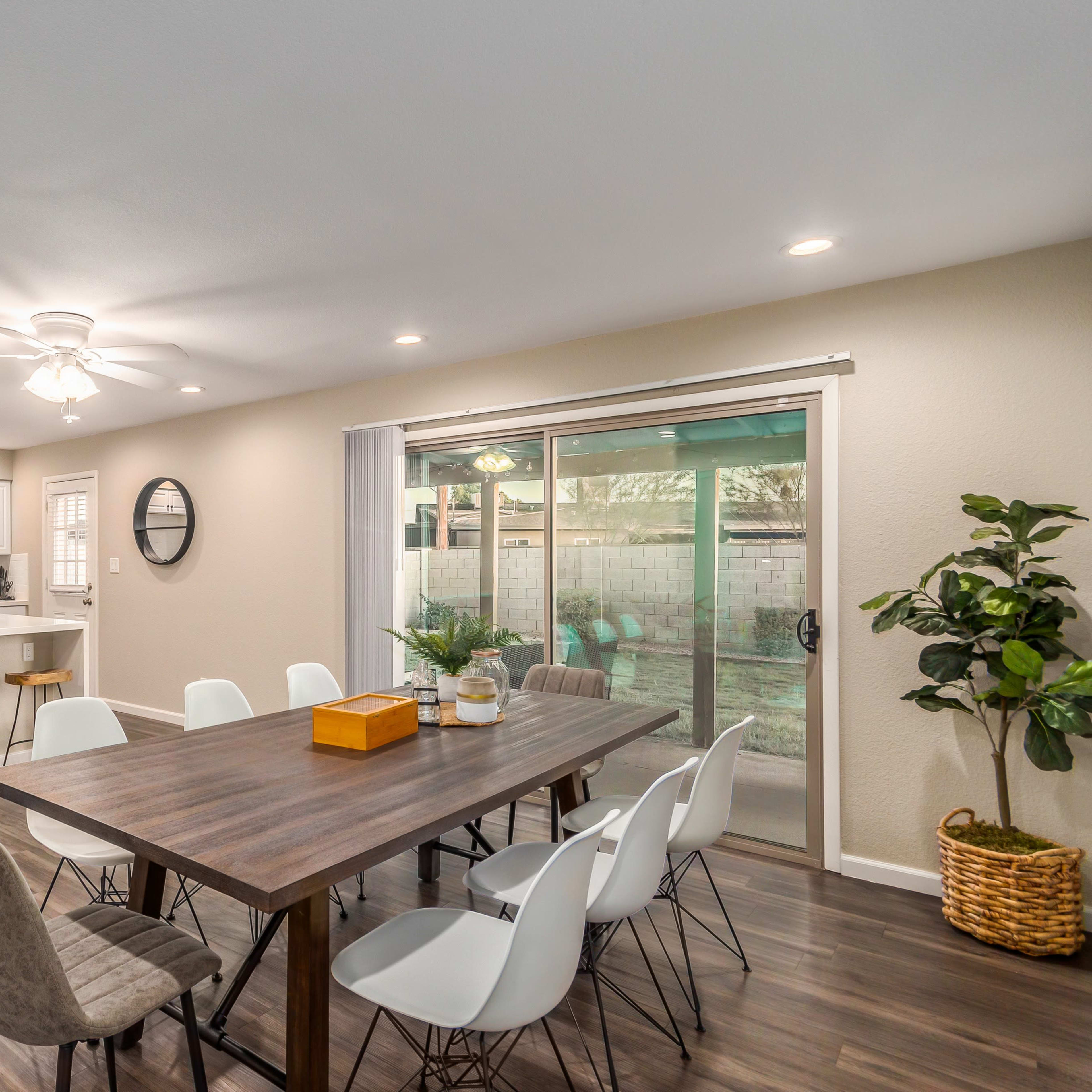 A modern dining area features a large wooden table surrounded by chairs, with an open kitchen and sliding glass doors leading to a patio.