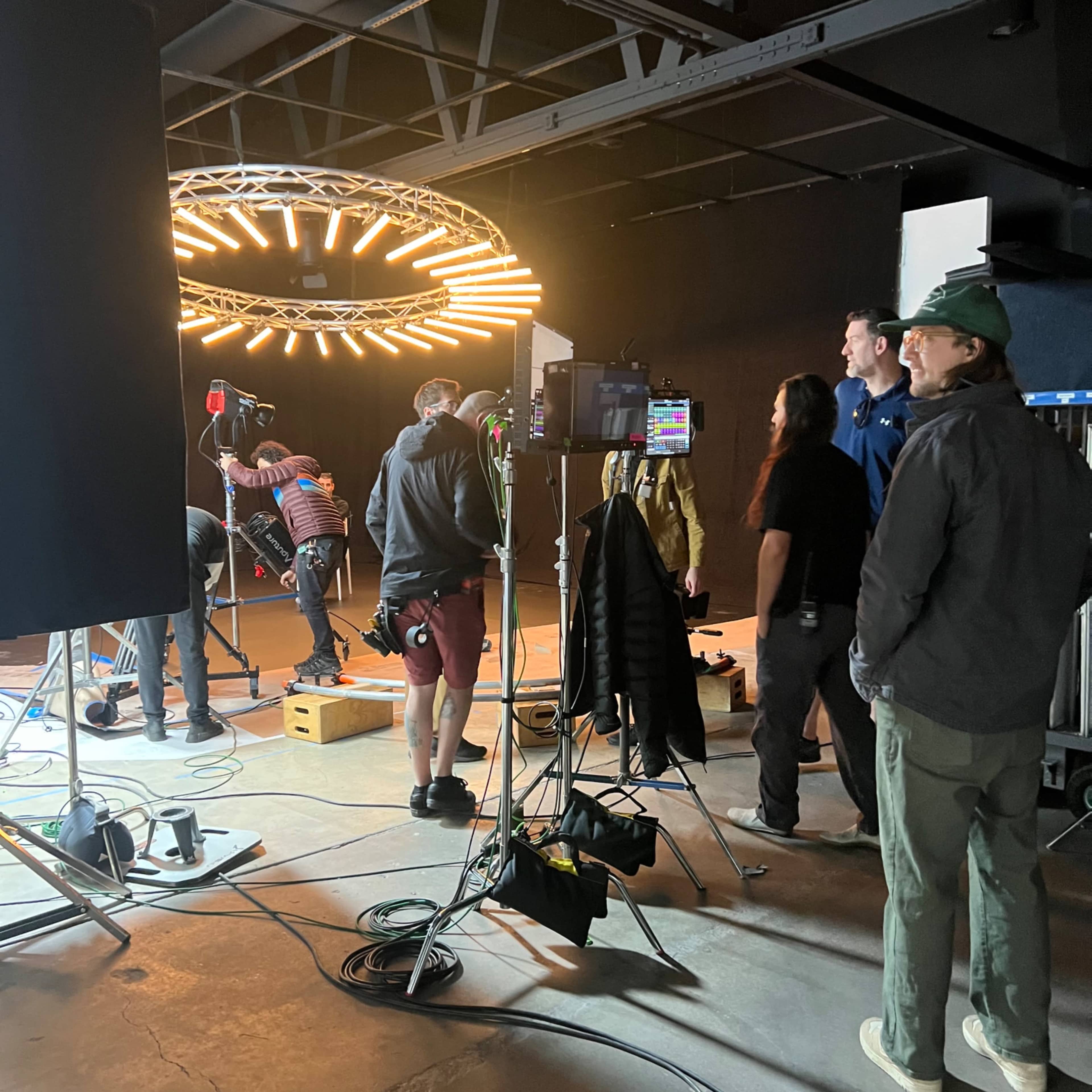 A film crew sets up equipment under a circular light fixture in a studio space.