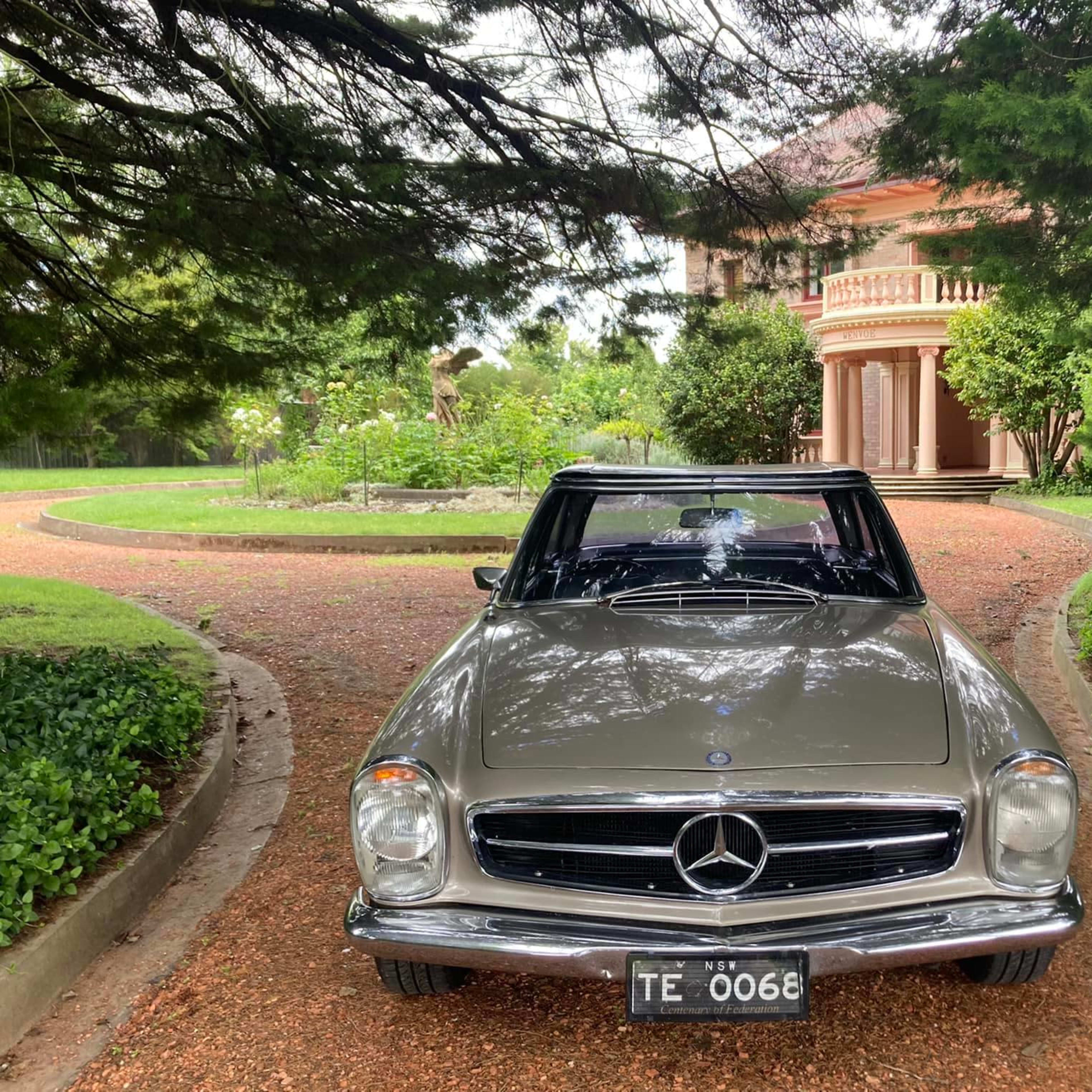 A vintage Mercedes-Benz is parked on a gravel driveway leading to a large, elegant house surrounded by lush greenery.
