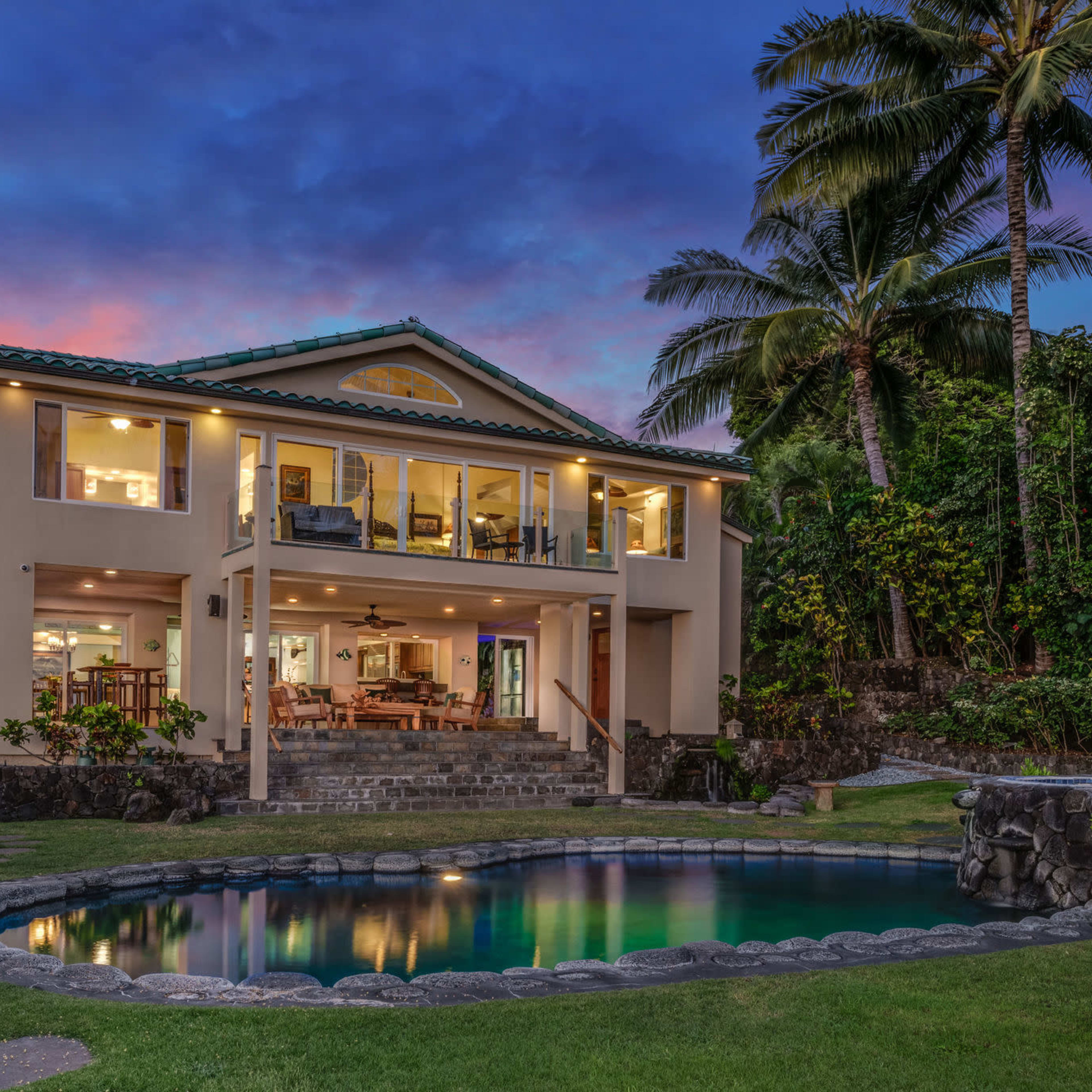A two-story house surrounded by tropical vegetation, with a swimming pool in the foreground and a colorful sky at dusk.