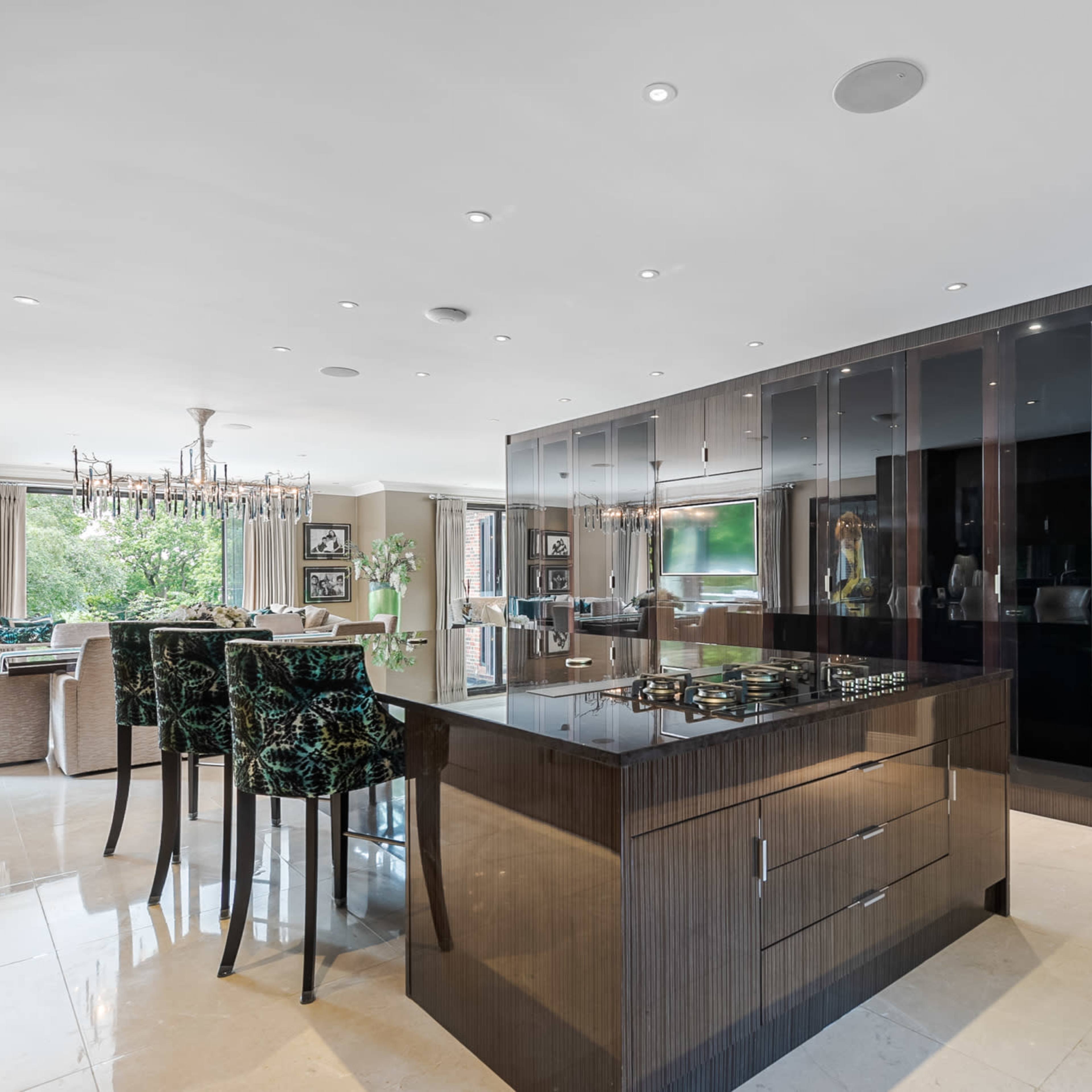 A modern kitchen features a central island with a stove, dark cabinetry, and a dining area with elegant lighting and green upholstered chairs.
