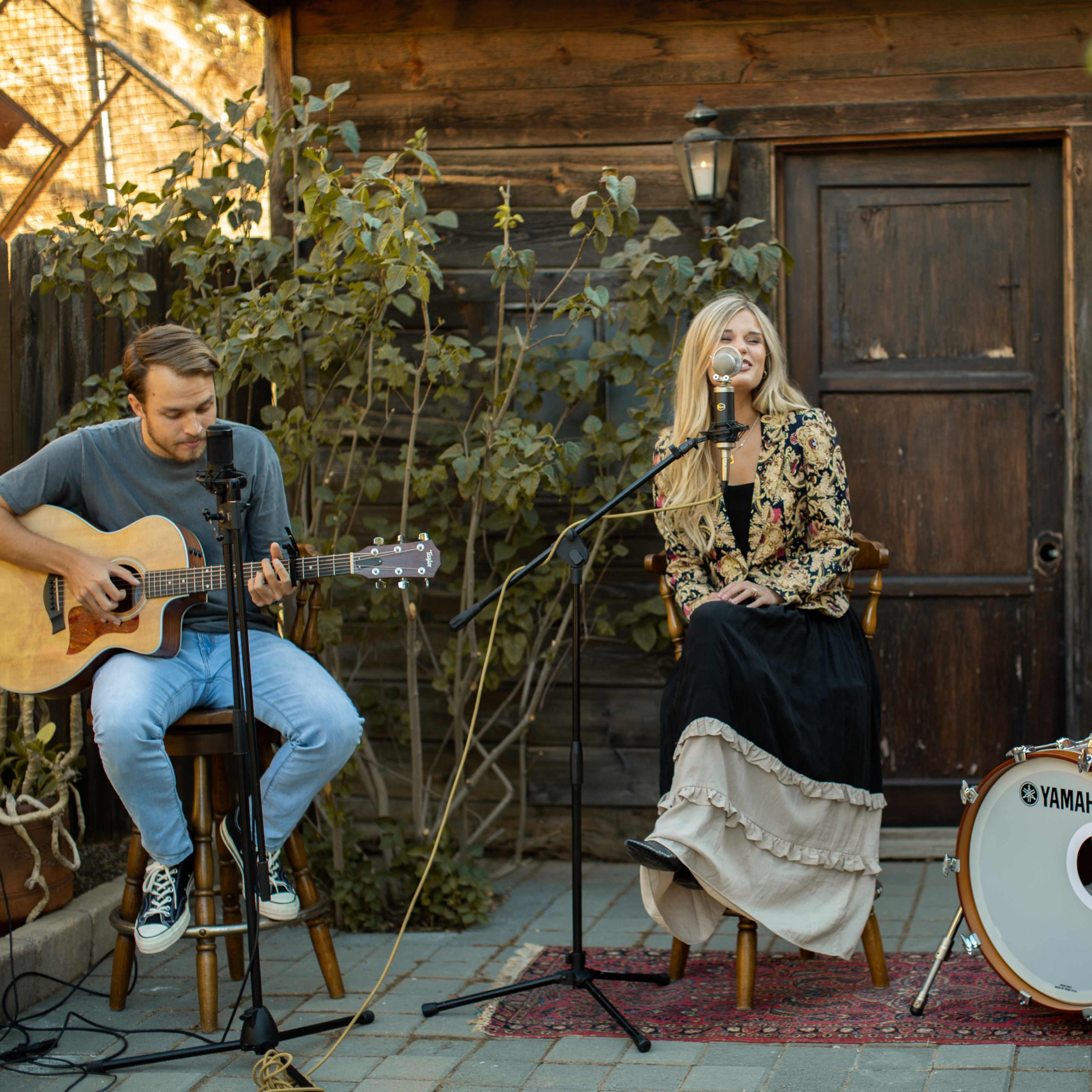 A musician plays guitar while a vocalist and a drummer perform in a garden setting.