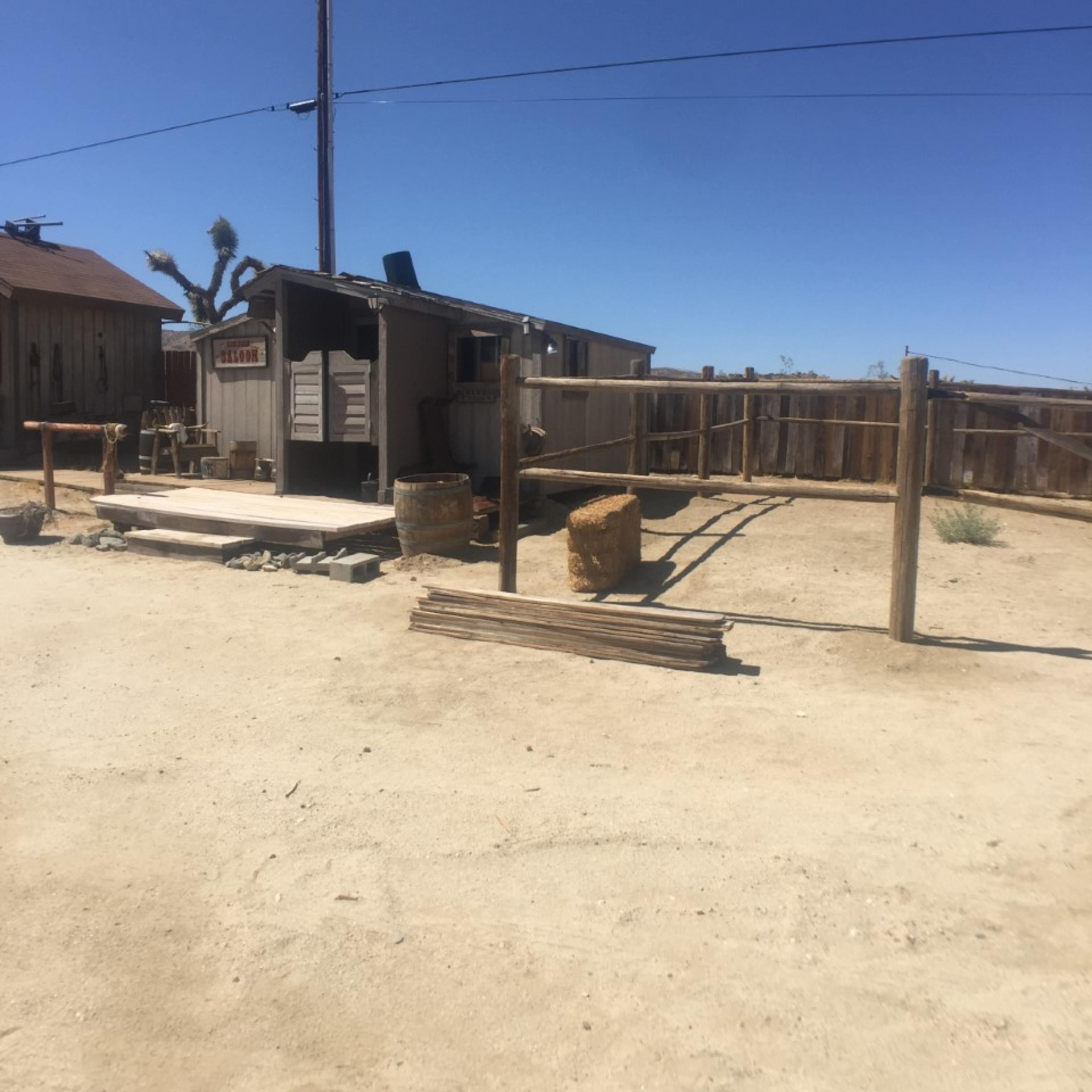 A small, wooden structure with a fenced enclosure sits on a sandy lot under a clear blue sky, surrounded by utility poles and sparse vegetation.