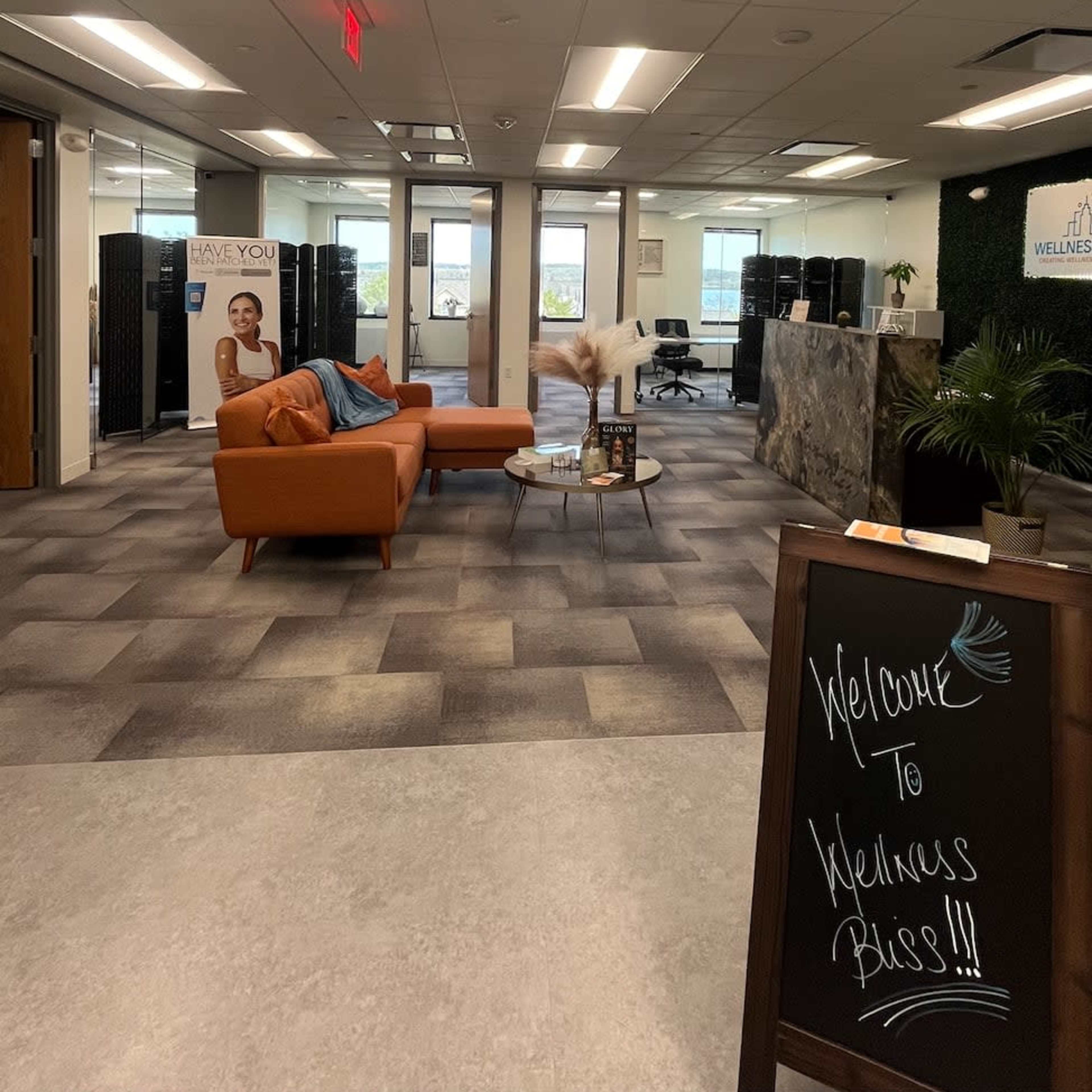 A modern office reception area with a seating arrangement featuring an orange couch, a welcome sign, and large windows offering a view outside.