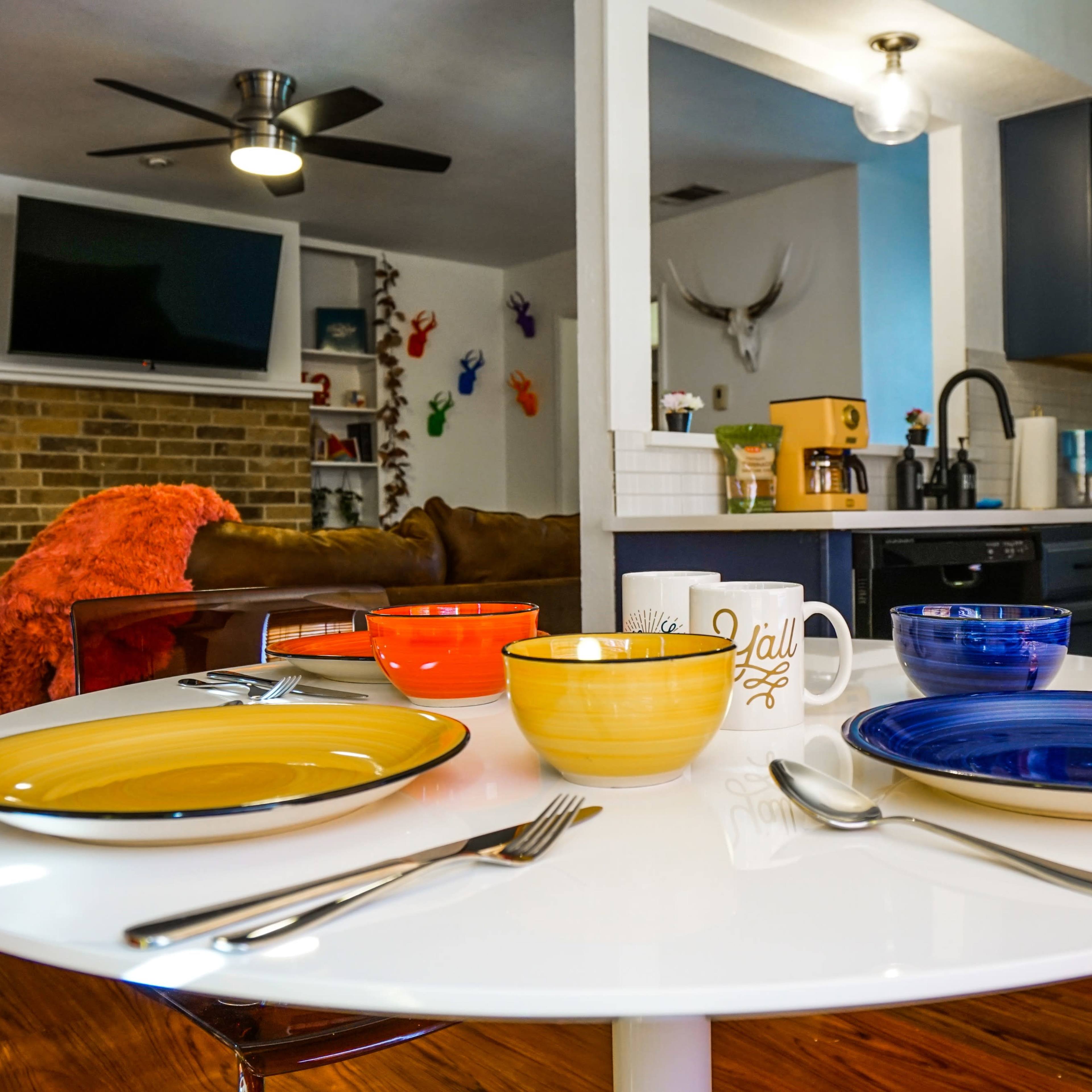 A round dining table is set with colorful plates and bowls, while a glimpse of a kitchen and living area can be seen in the background.