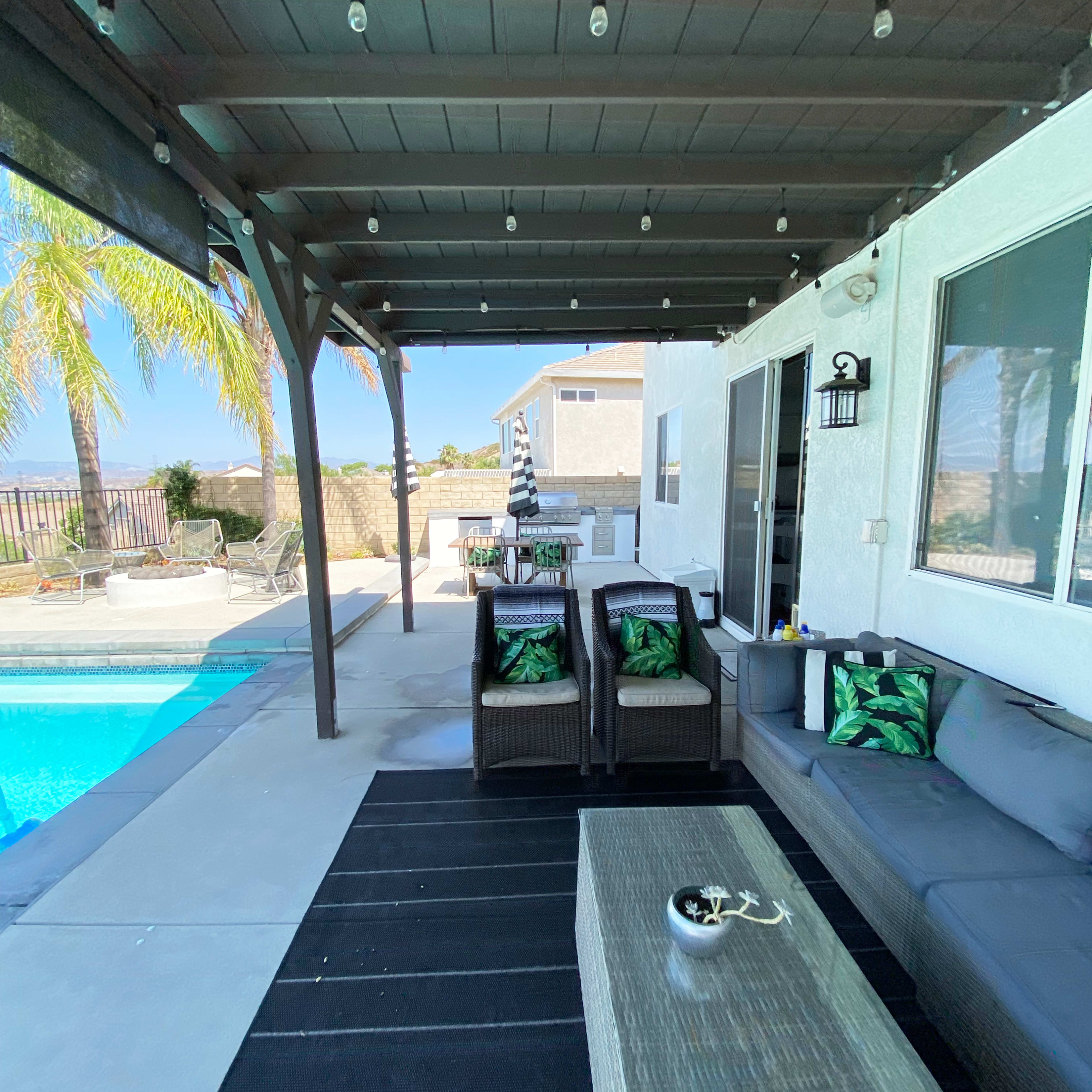 A covered patio features seating and a coffee table beside a swimming pool with palm trees in the background.