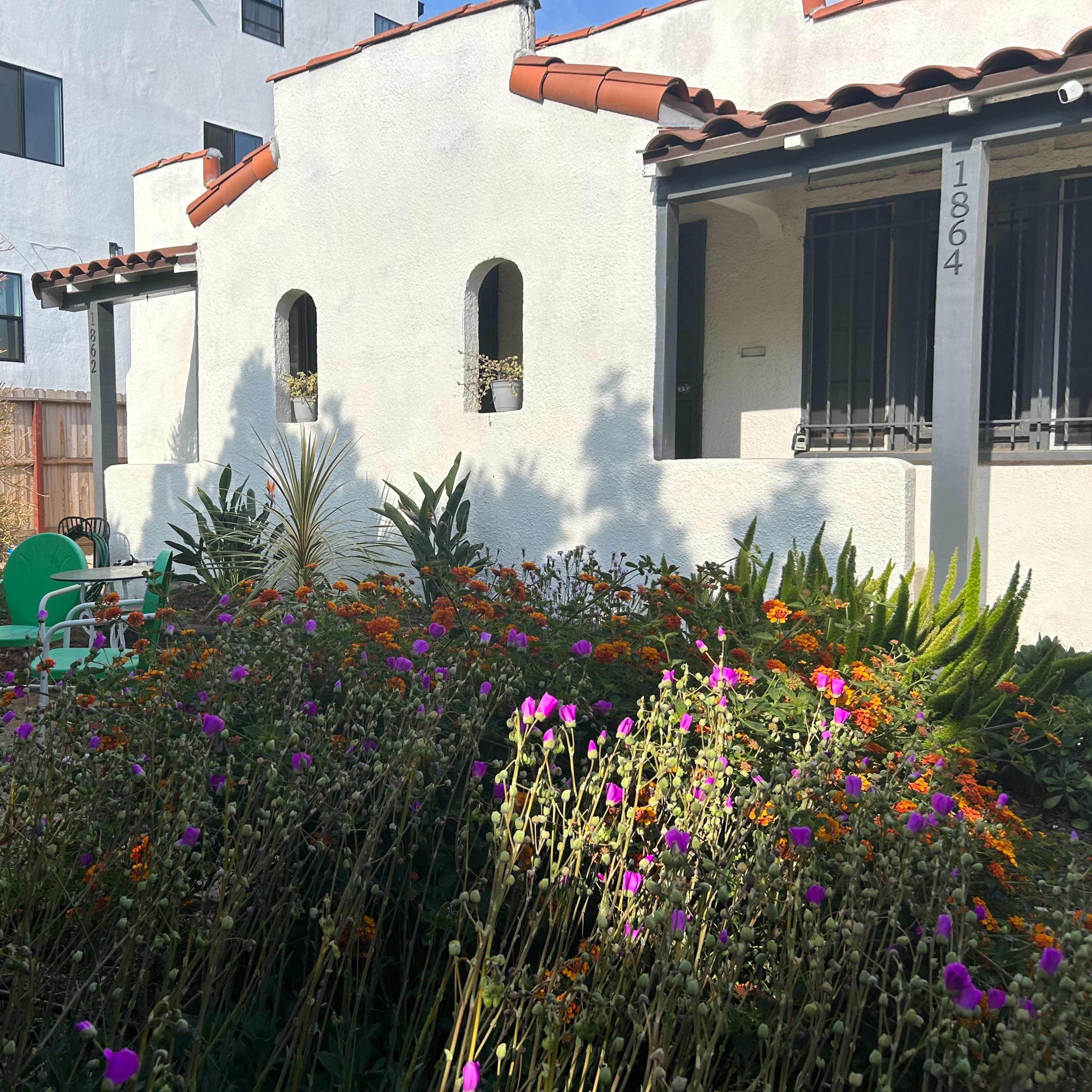 A small garden filled with vibrant flowers and plants surrounds a white stucco building with a tiled roof and visible windows.