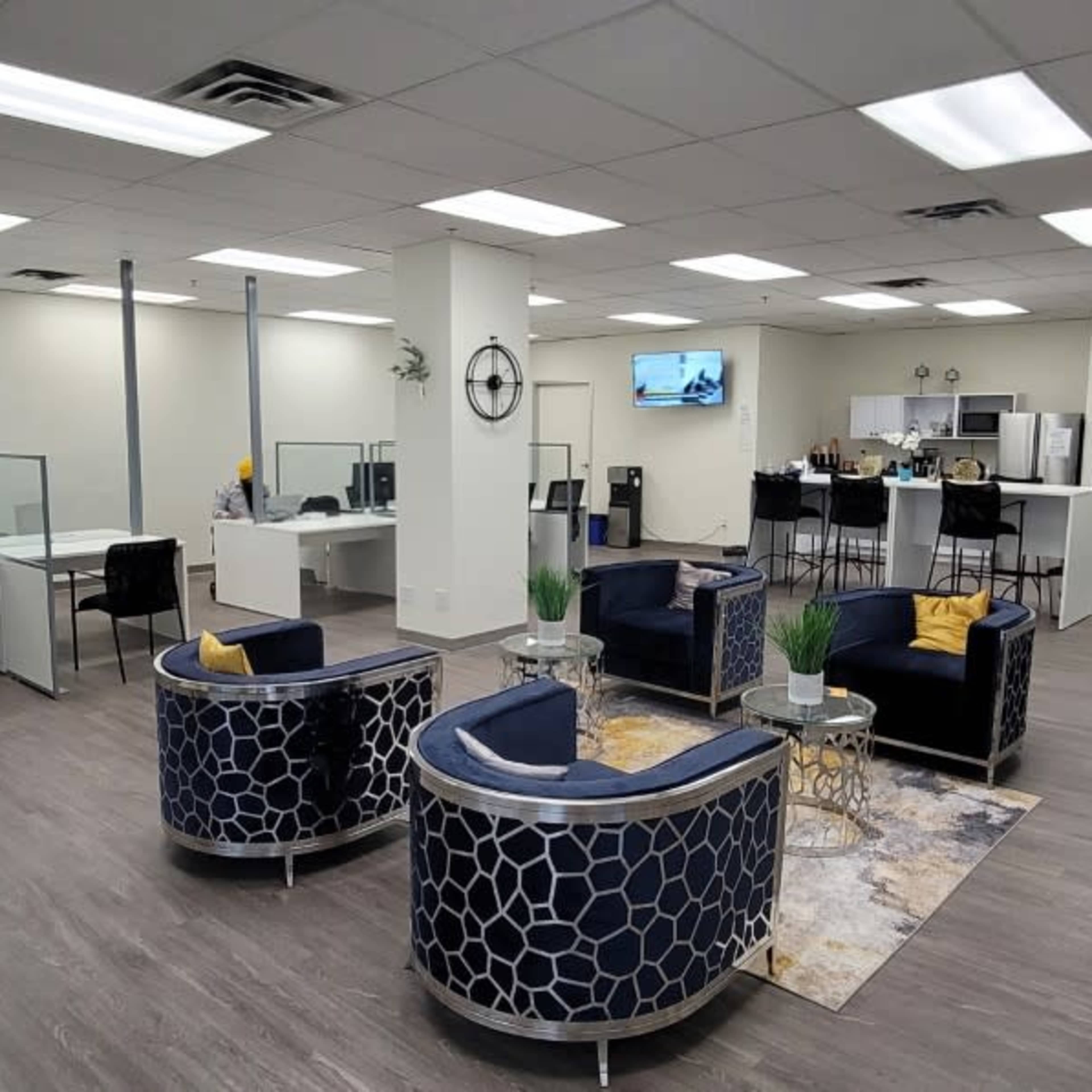 A modern office space with several workstations, a lounge area featuring two chairs, and a kitchenette visible in the background.