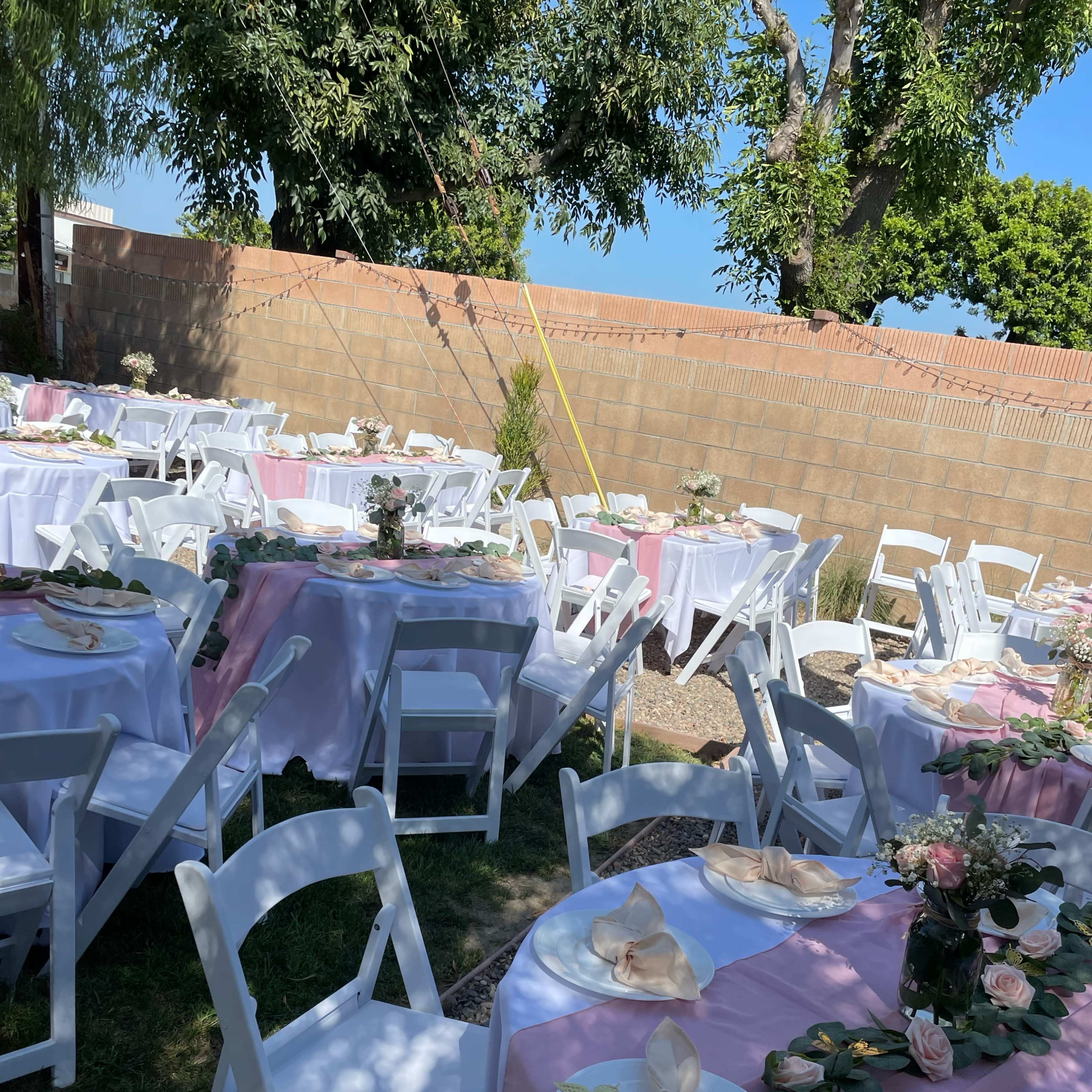 The scene shows a backyard setup for an event with multiple tables covered in pink and white tablecloths, surrounded by chairs and greenery.