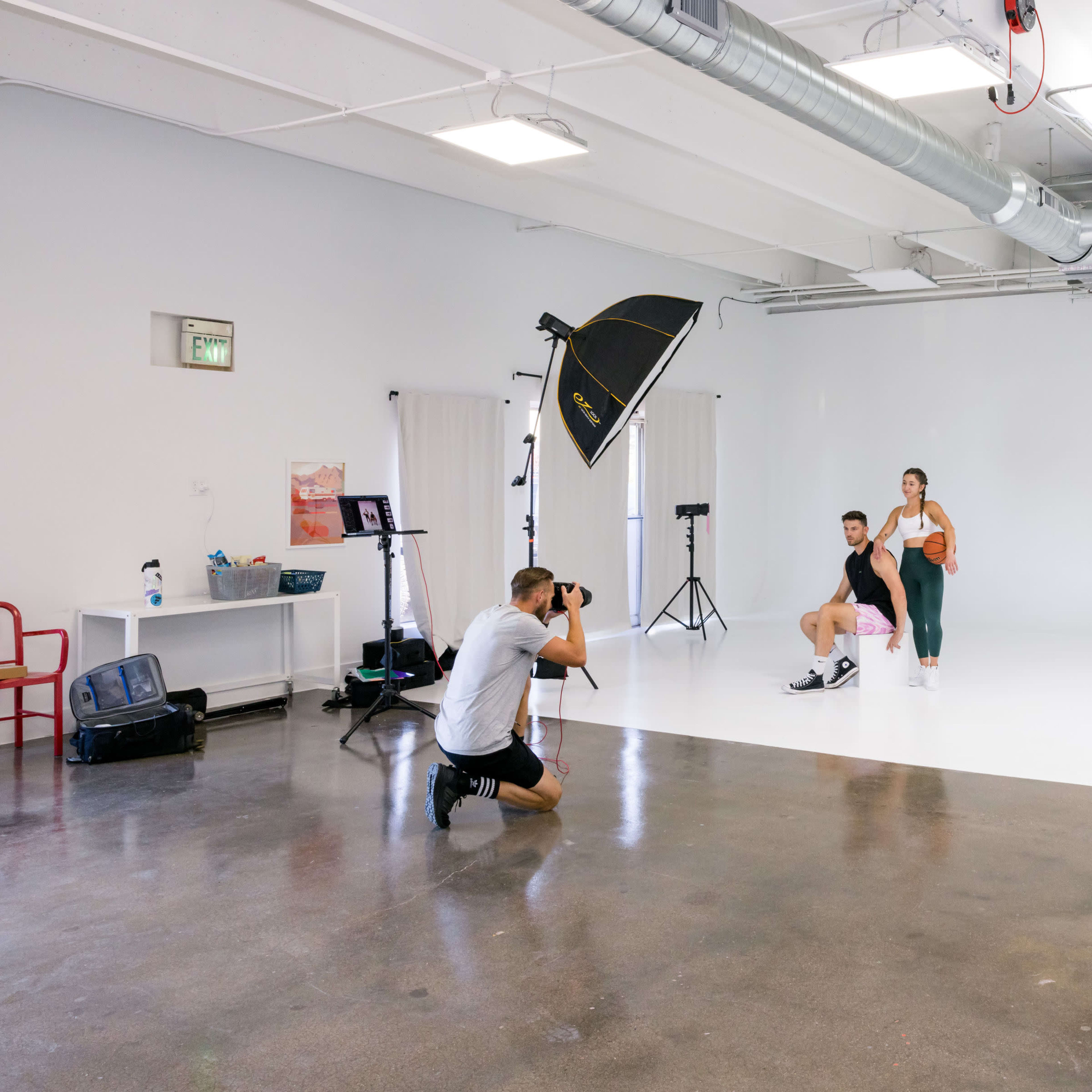 A photographer captures a male and female model in a studio setting with a white backdrop and various lighting equipment.