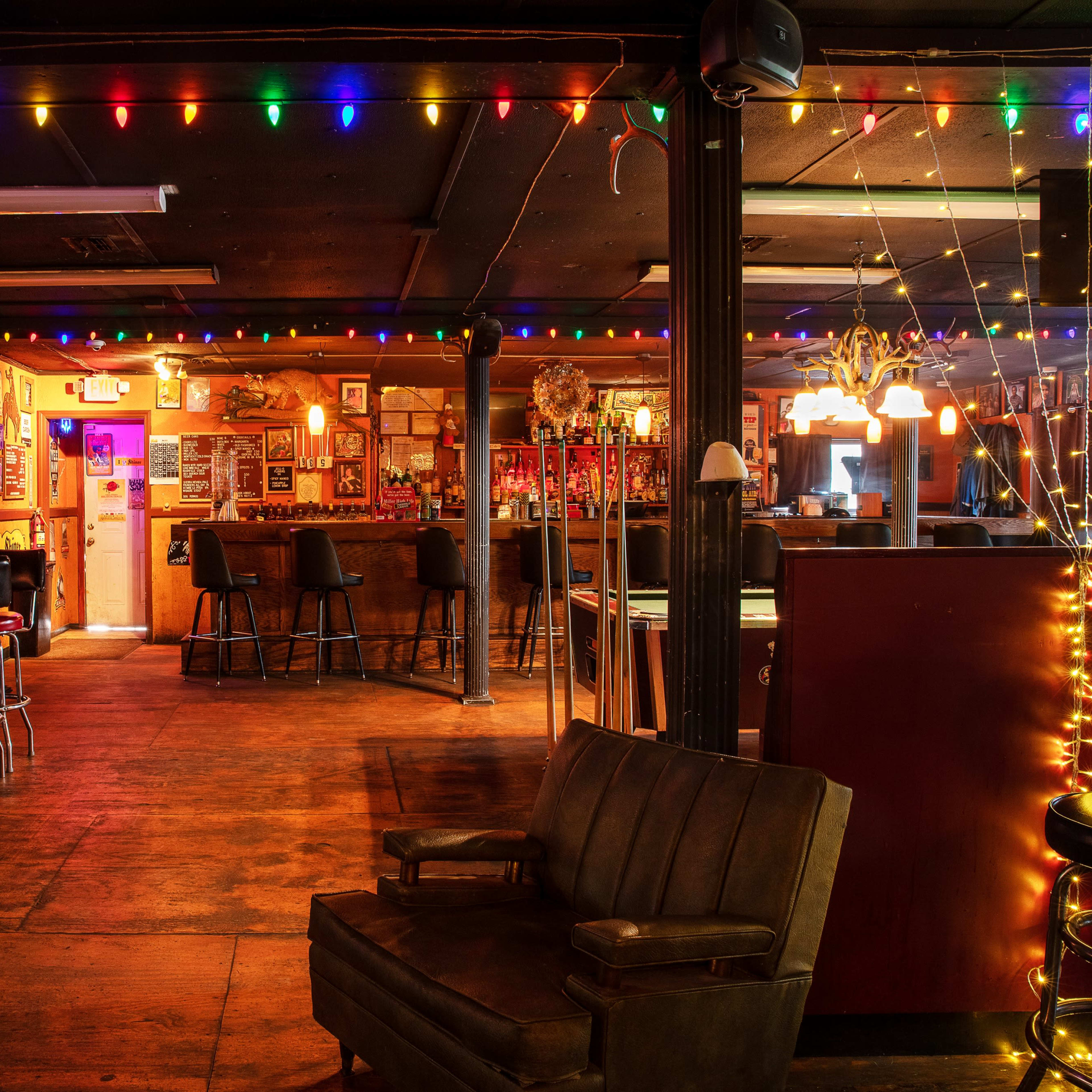 The interior of a dimly lit bar features colorful string lights, wooden floors, and a seating area with stools and a couch.