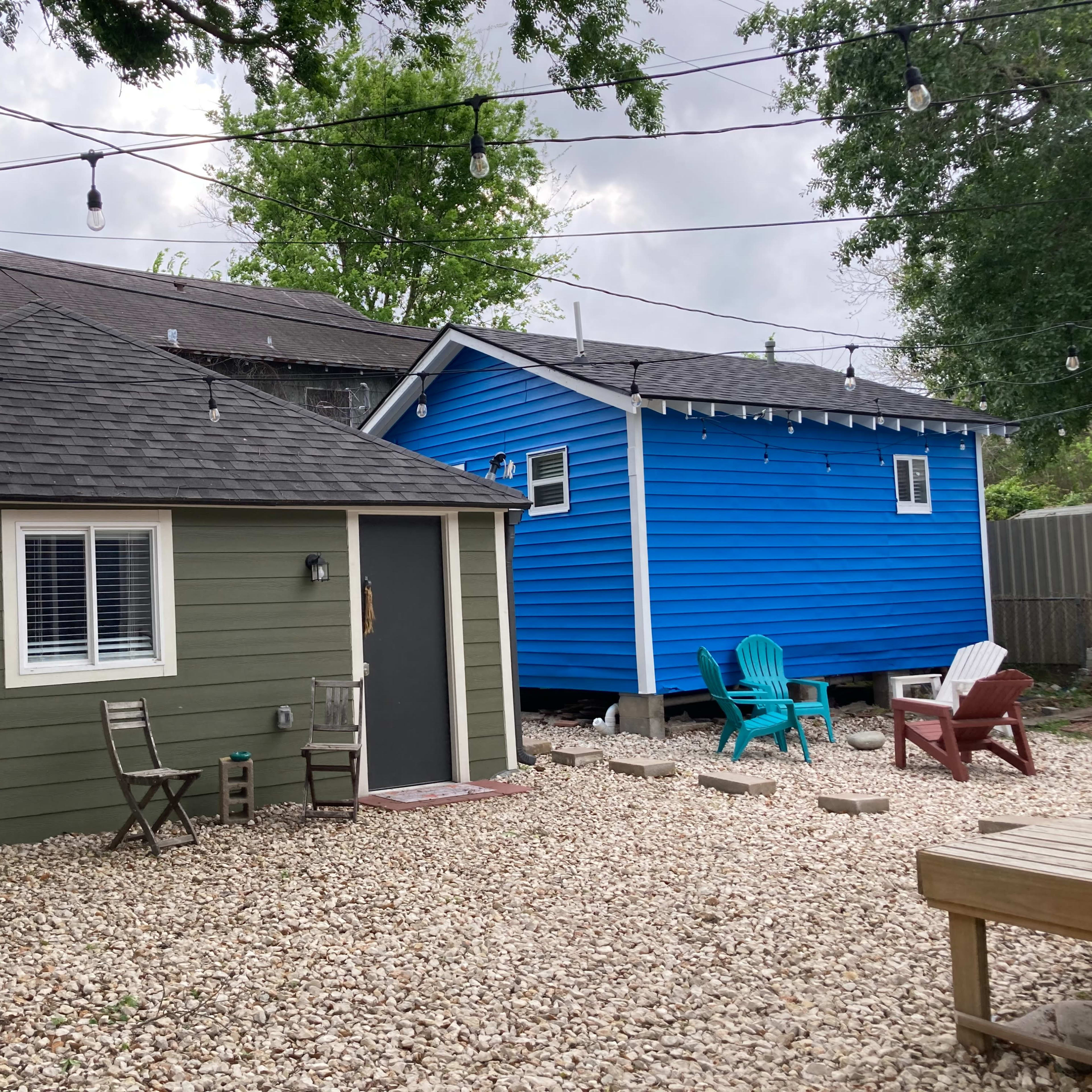 Two houses, one green and one blue, situated next to each other in a gravel yard with outdoor seating and string lights above.