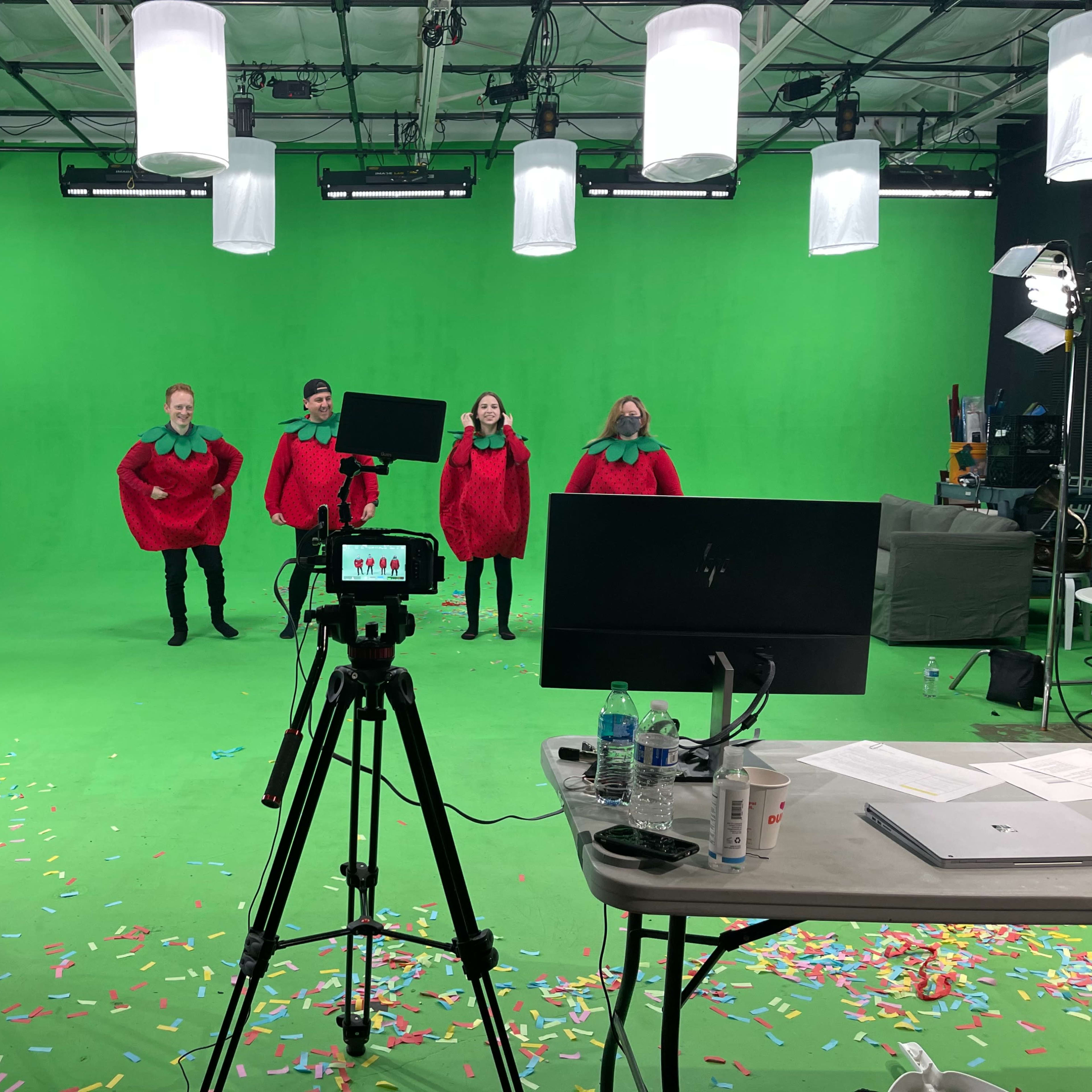A group of four individuals dressed in red costumes stands in front of a green screen, with a camera setup and colorful confetti scattered on the floor.
