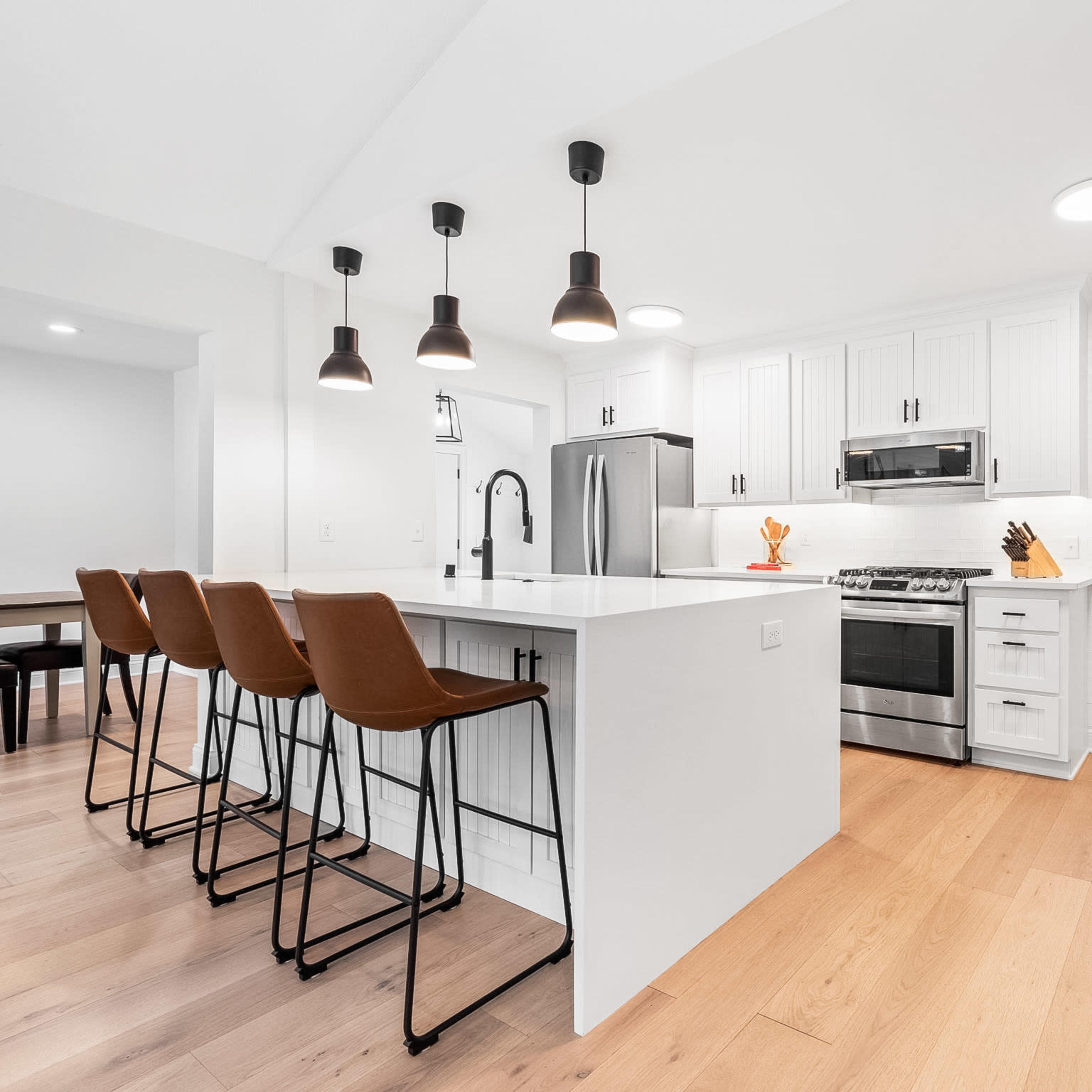A modern kitchen featuring an island with three bar stools, white cabinetry, stainless steel appliances, and pendant lighting.